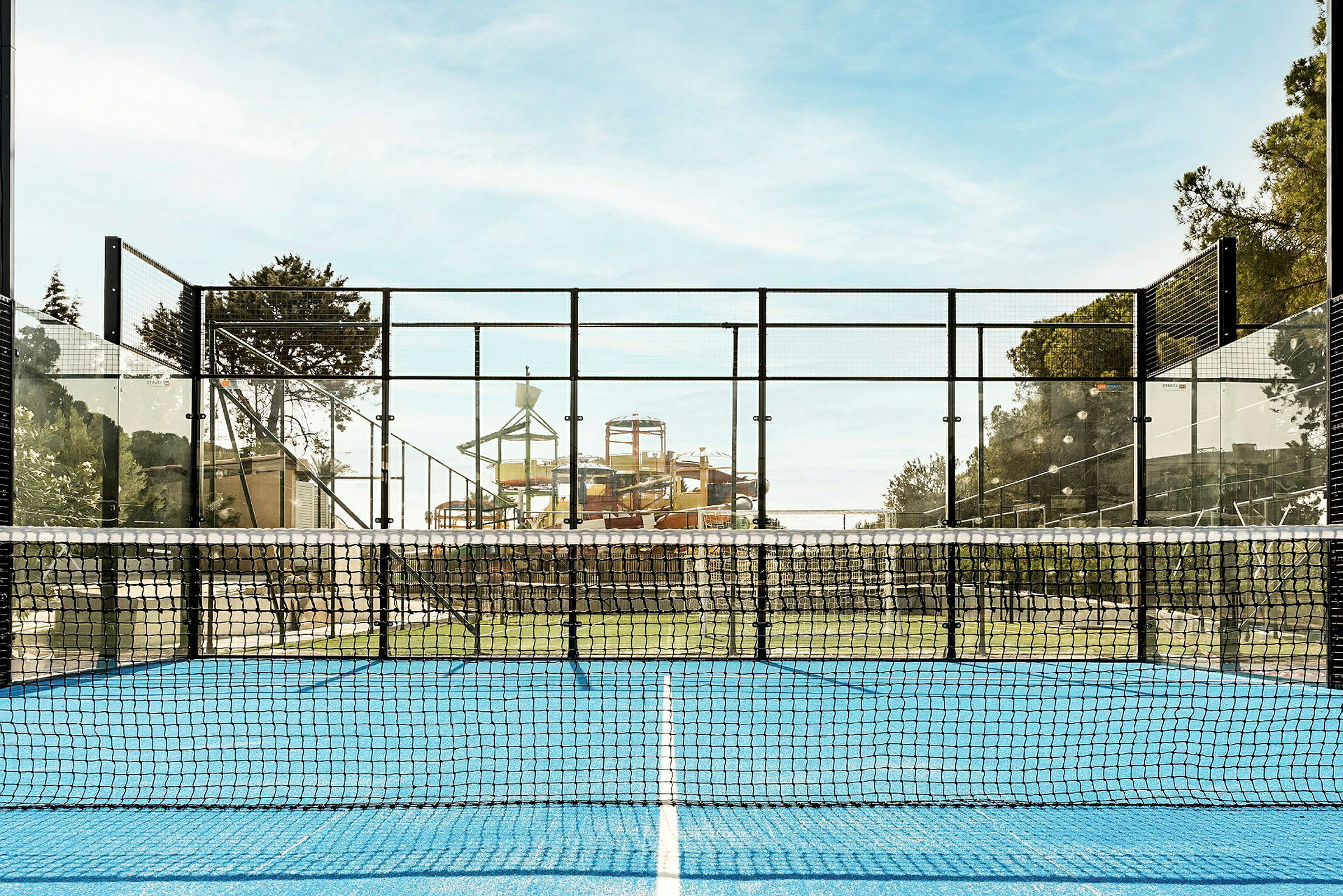 En padelbana med blått underlag och nät i förgrunden, med en vattenpark i bakgrunden. (AI-genererad text)