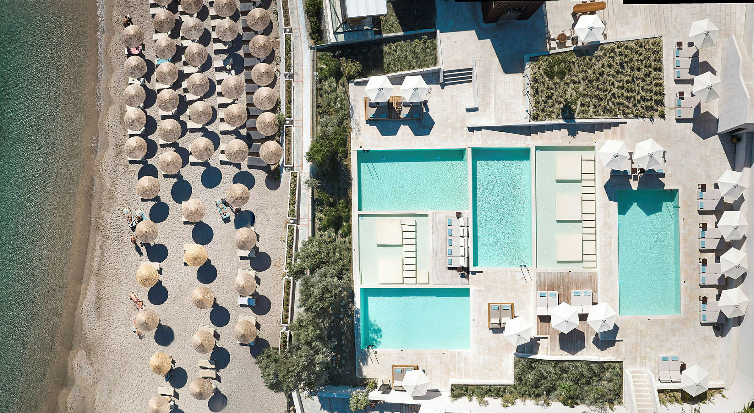 Flygvy över en strand med parasoller och solstolar bredvid en anläggning med flera swimmingpooler. (AI-genererad text)