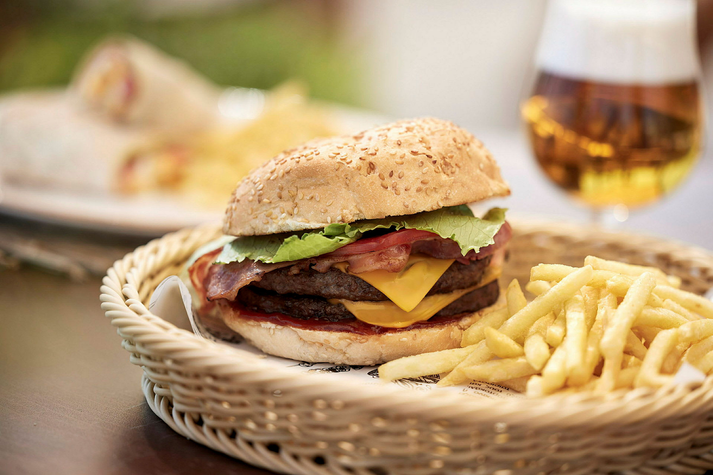 Hamburgare med ost och bacon, serverad med pommes frites på en korg, samt en öl i bakgrunden. (AI-genererad text)