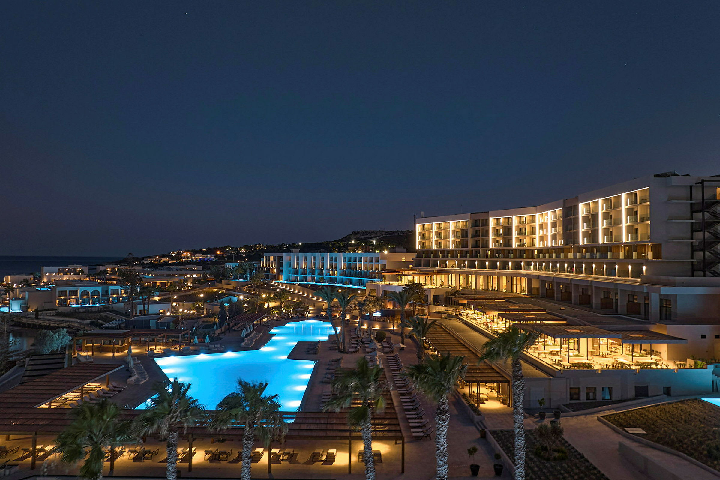 Nattvy av ett upplyst hotell med poolområde och palmer. (AI-genererad text)
