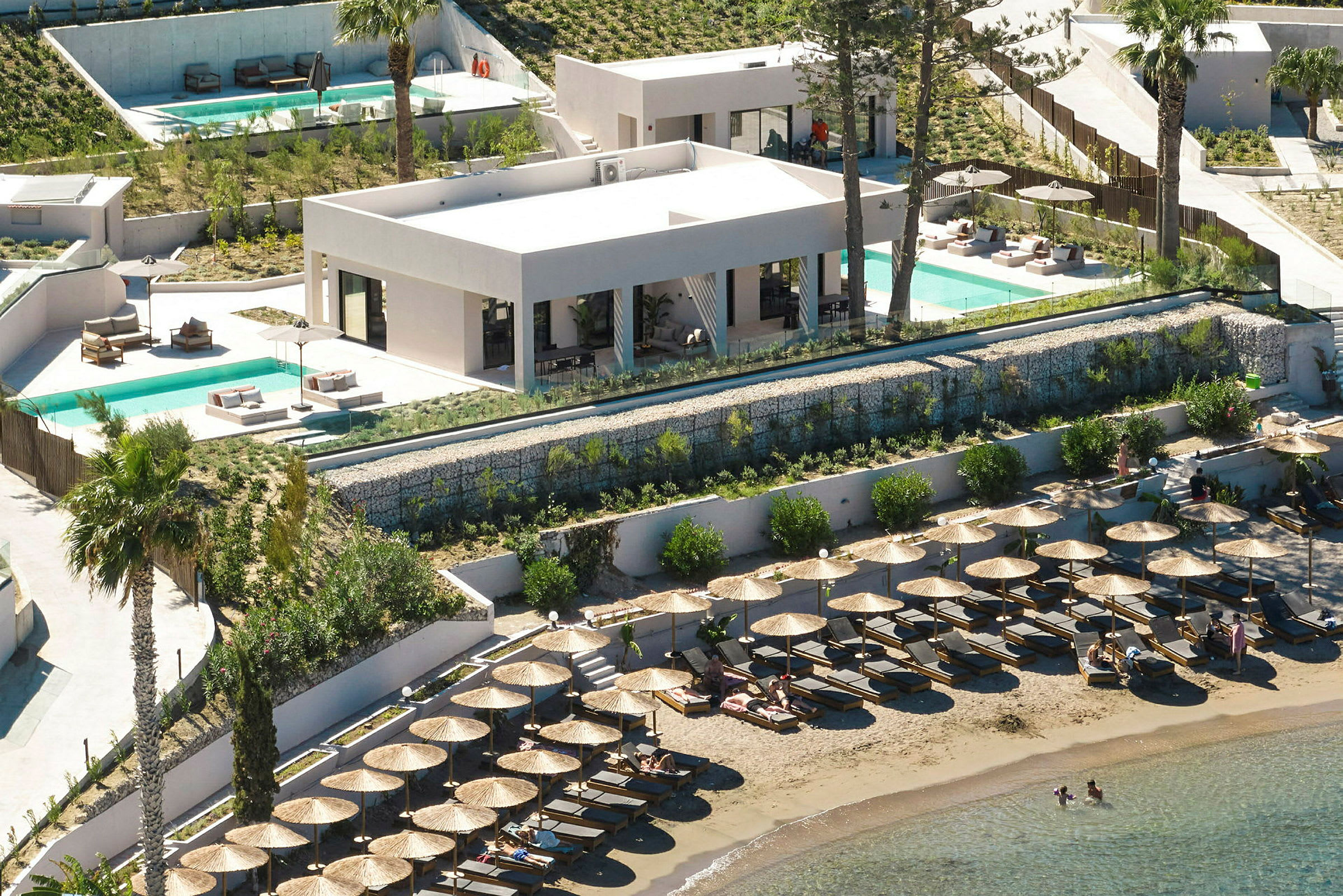 Utsikt över en strand med solstolar och parasoller, bakom en byggnad med pool och terrass. (AI-genererad text)