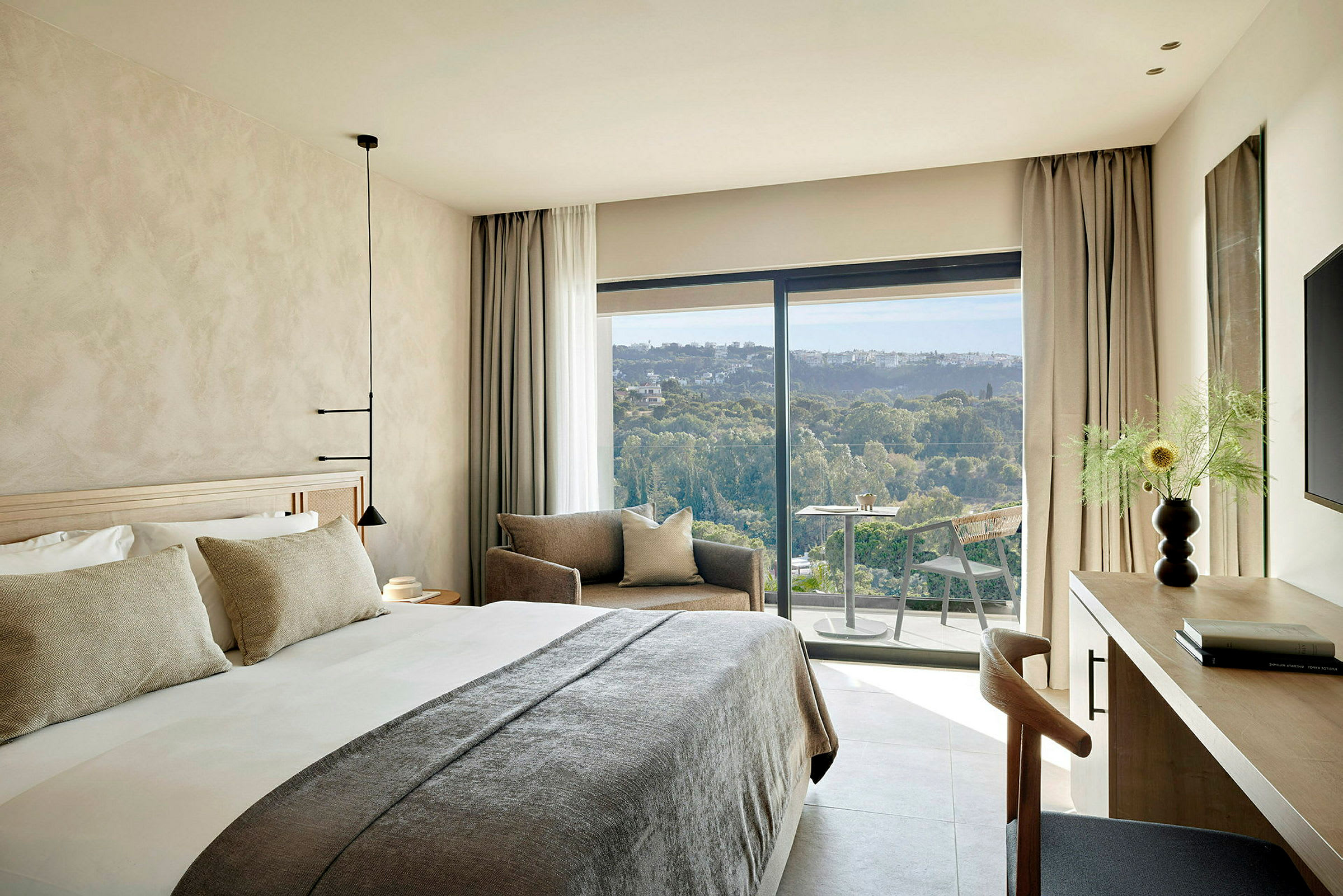 Ett modernt hotellrum med en stor säng, sittgrupp och en balkong med utsikt över ett grönt landskap. (AI-genererad text)