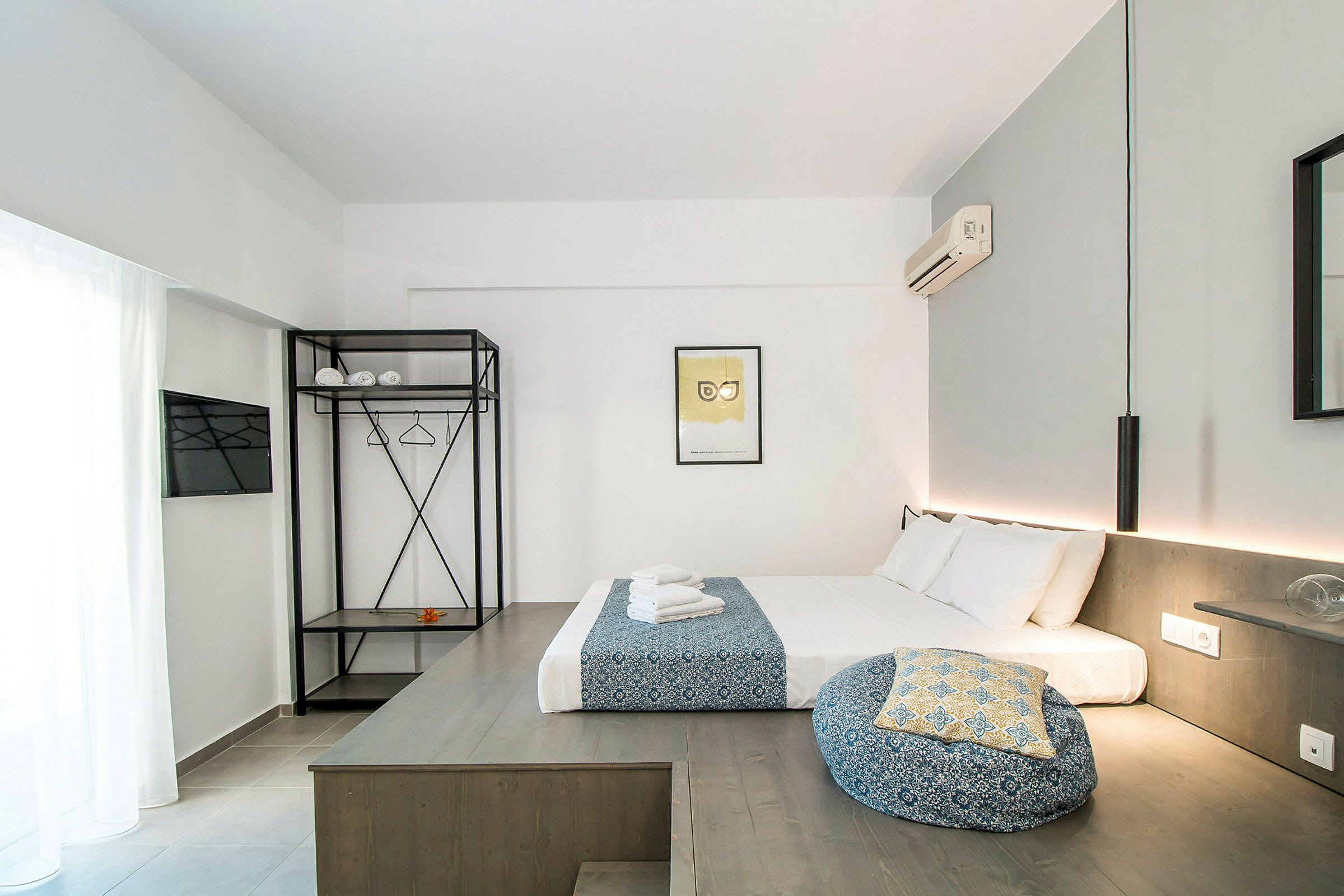 Ett modernt hotellrum med en säng bäddad med vita och blåfärgade textilier, en öppet garderob med hyllplan och klädhängare samt en väggmonterad luftkonditionering. (AI-genererad text)