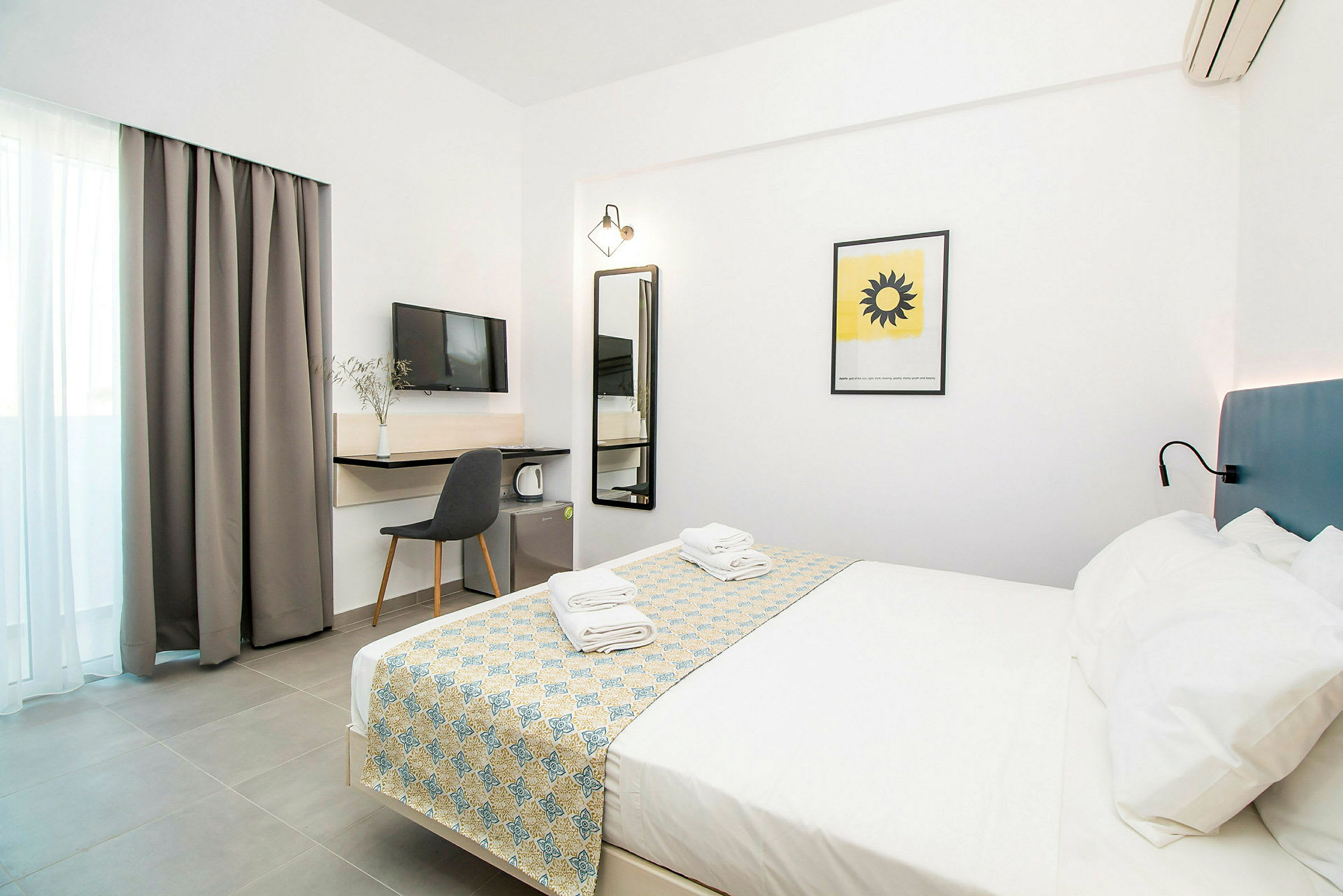 Ett hotellrum med vit inredning, en dubbelsäng bäddad med mönstrat överkast och handdukar, samt möbler och en TV. (AI-genererad text)
