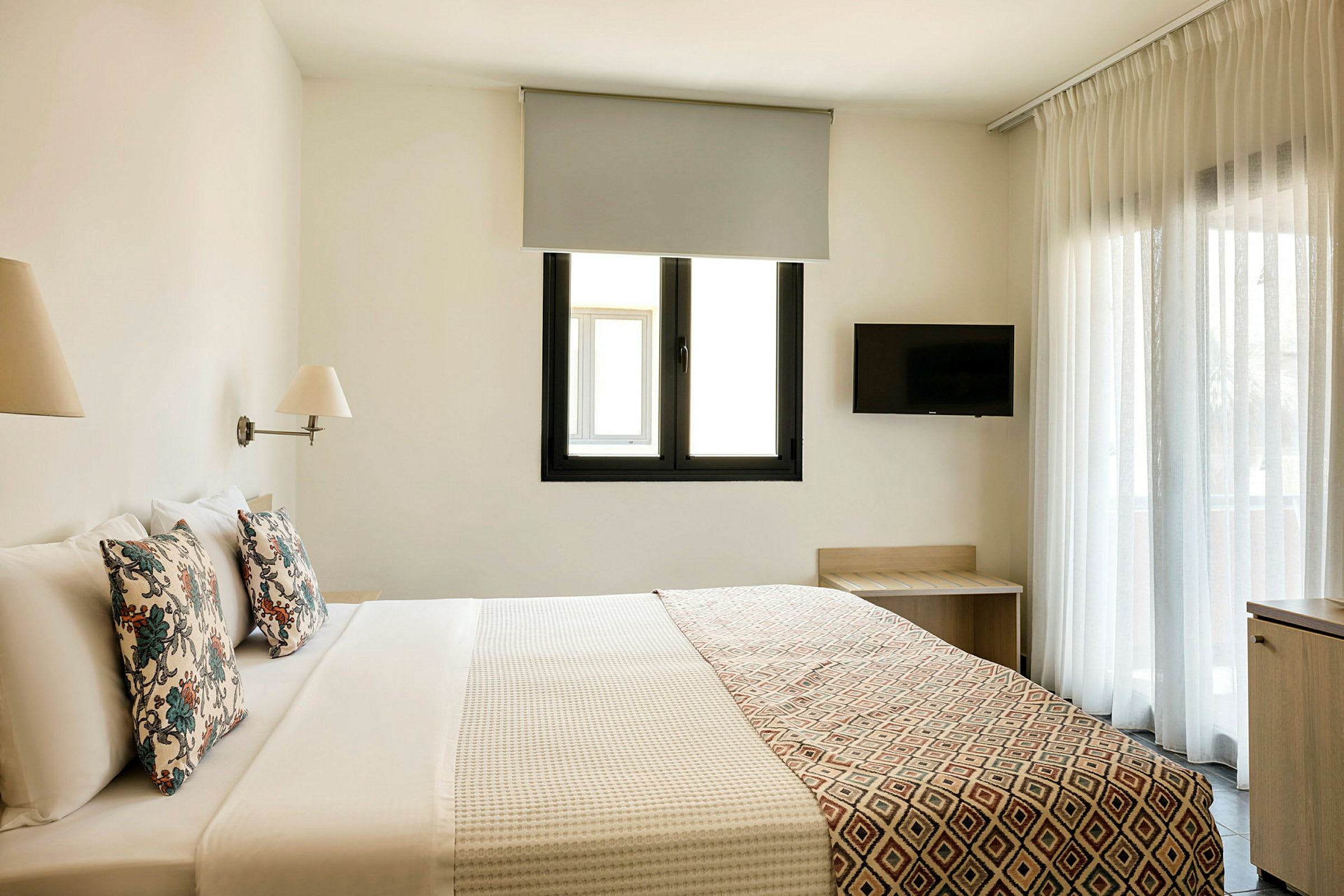 Ett hotellrum med en dubbelsäng, två lampor, en tv på väggen och ett fönster med gardiner. (AI-genererad text)