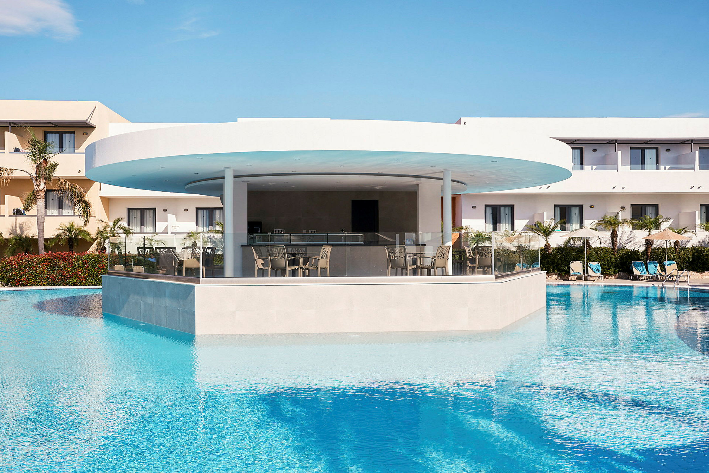 En modern hotellbar omgiven av en pool med klart vatten och solstolar i bakgrunden. (AI-genererad text)