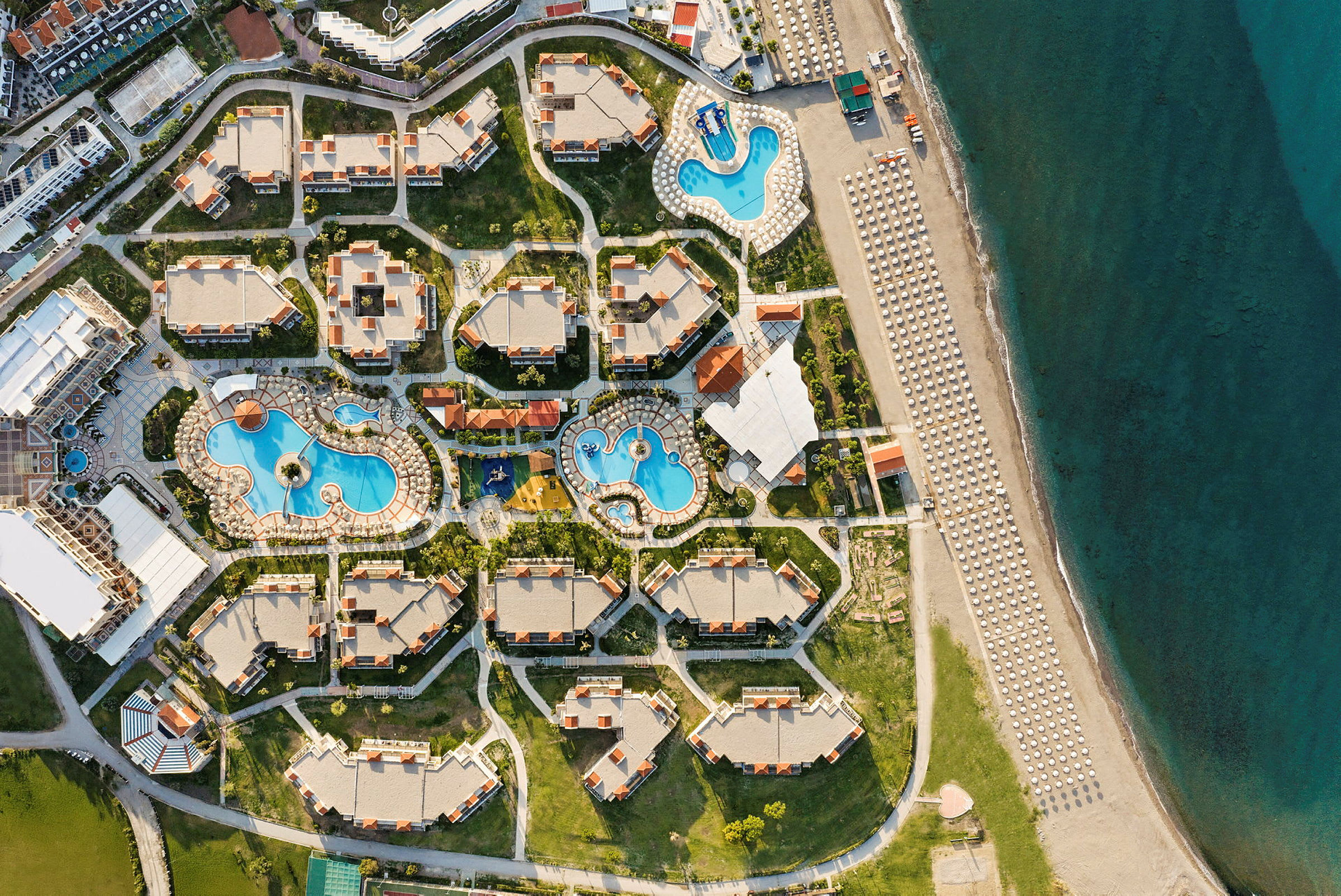Flygbild av en semesteranläggning med pooler, byggnader och närliggande strand. (AI-genererad text)