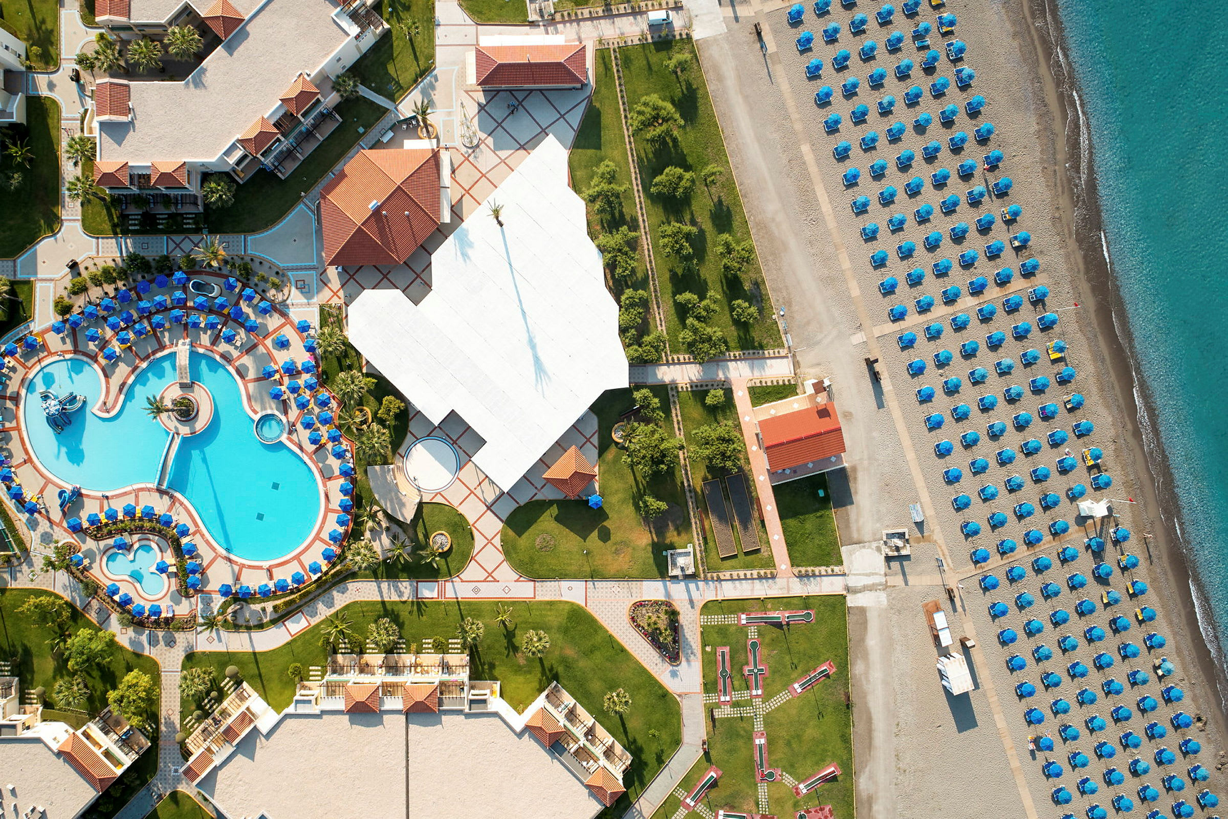 Flygbild som visar ett semesterhotell med poolområde, grönområden och en strand med uppställda parasoller. (AI-genererad text)