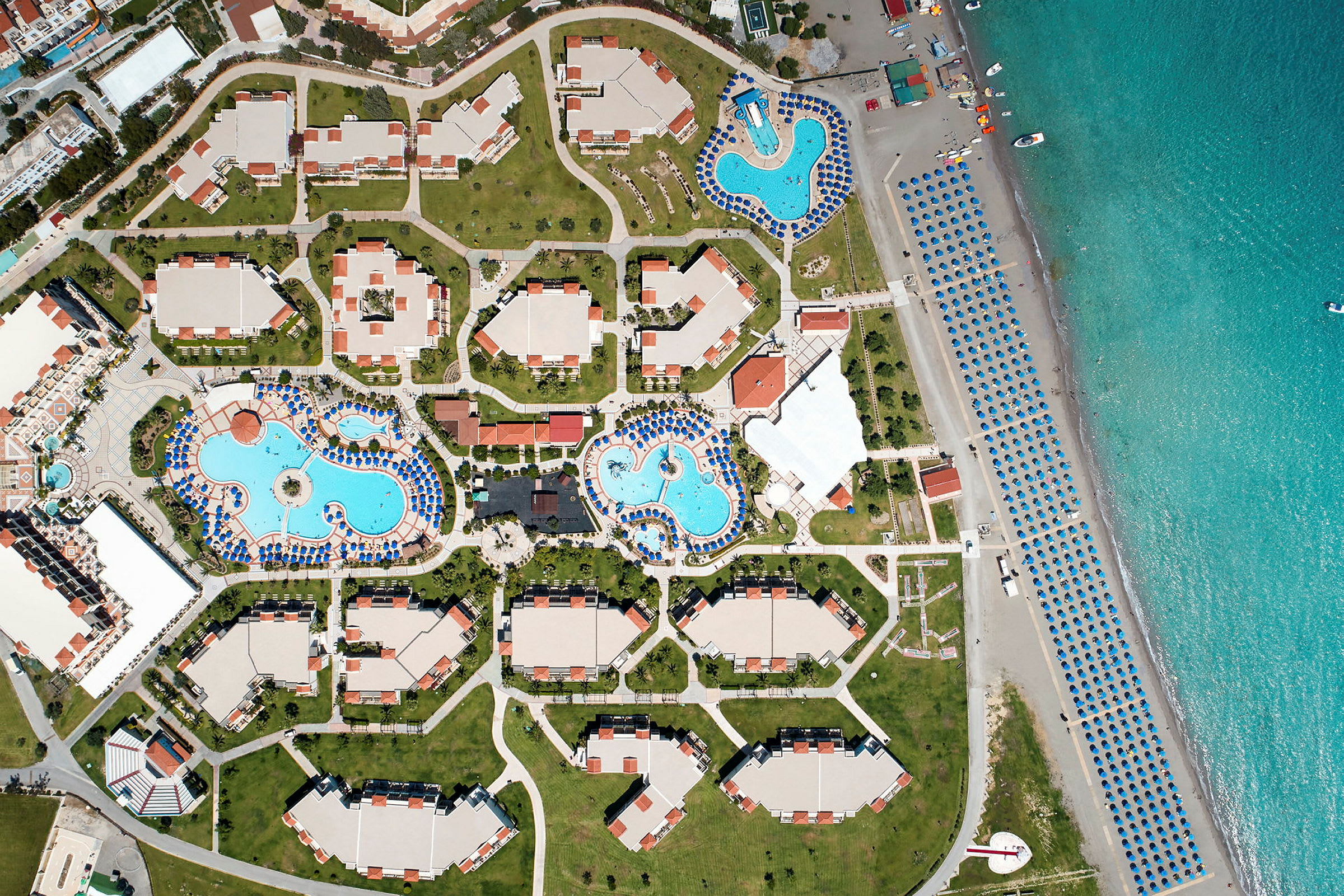 Fågelperspektiv av en resort med flera byggnader, poolområden och strand med parasoller. (AI-genererad text)