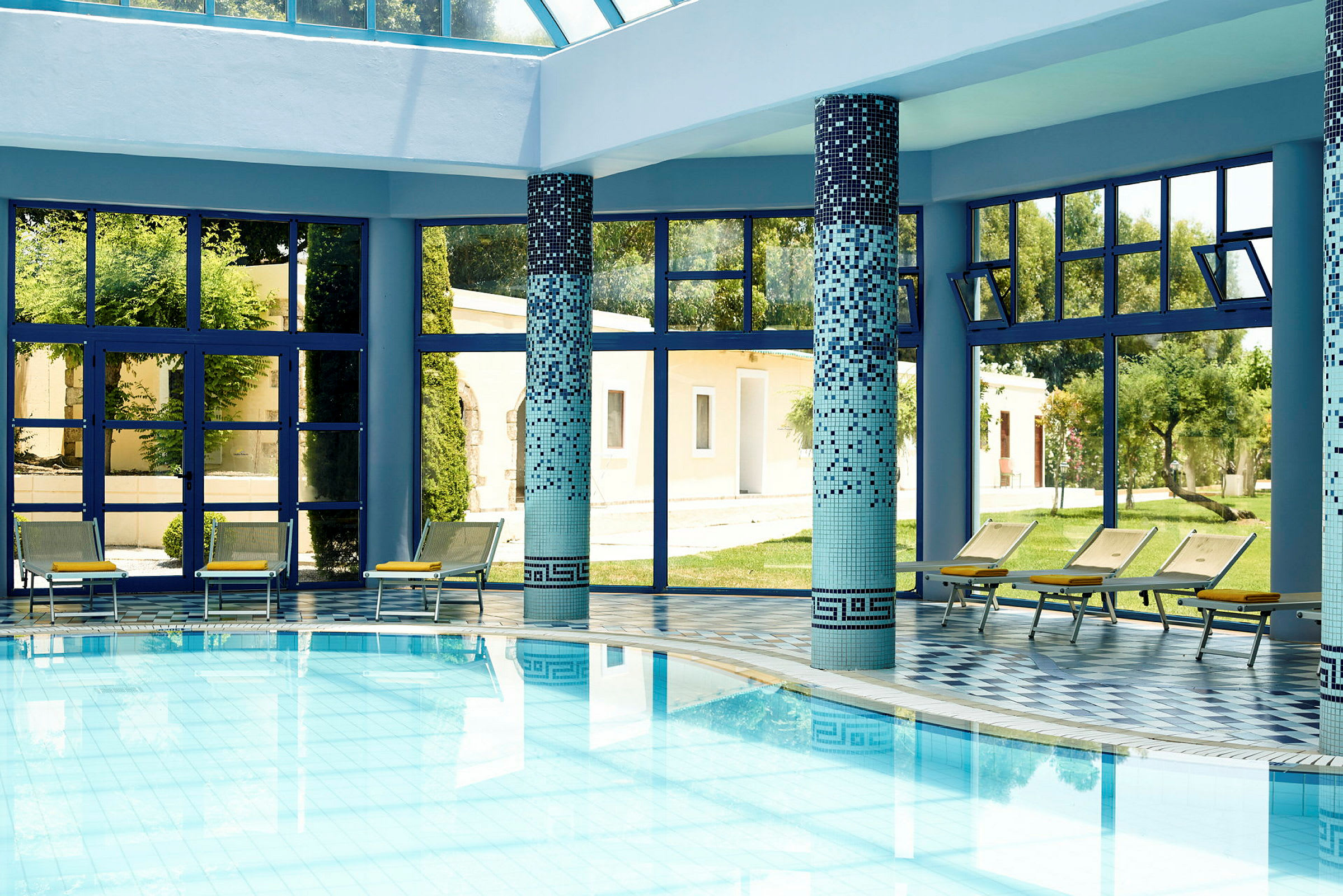 Inomhuspool med blå mosaik och solstolar vid fönster. (AI-genererad text)