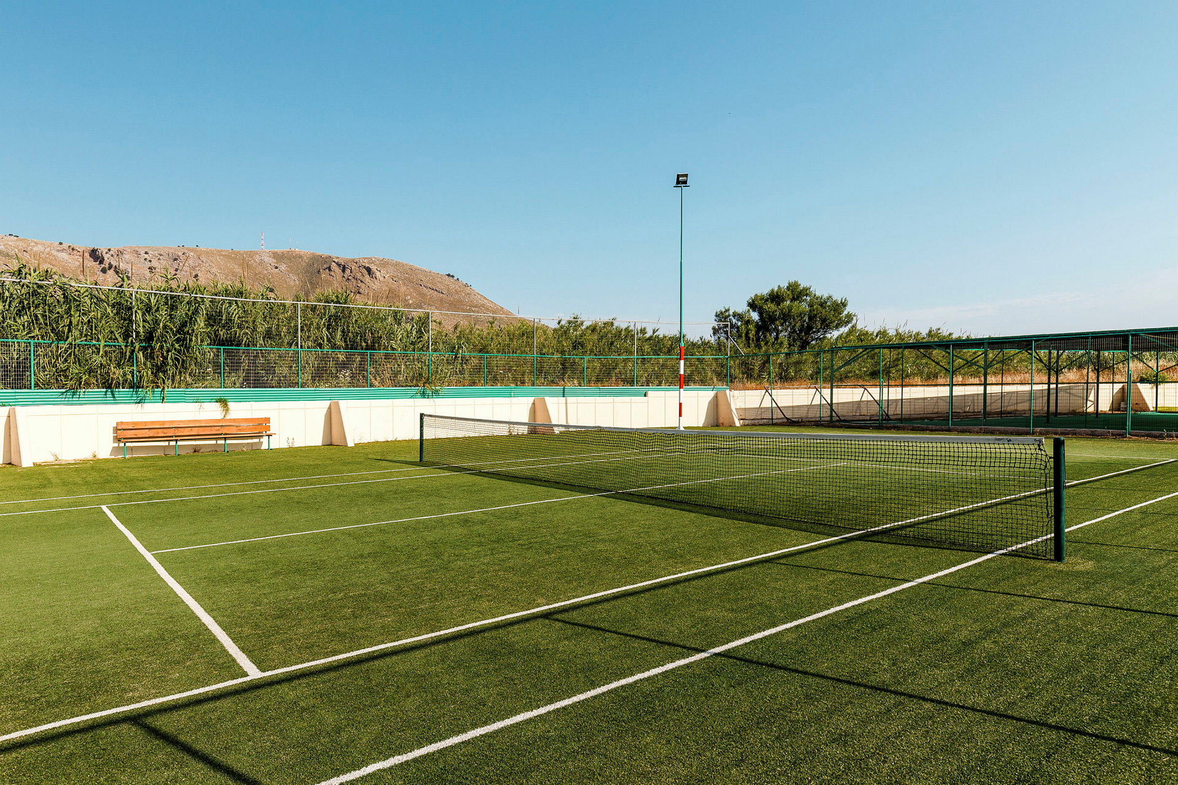 En tennisbana med grön konstgräs, omgiven av staket och vegetation. (AI-genererad text)