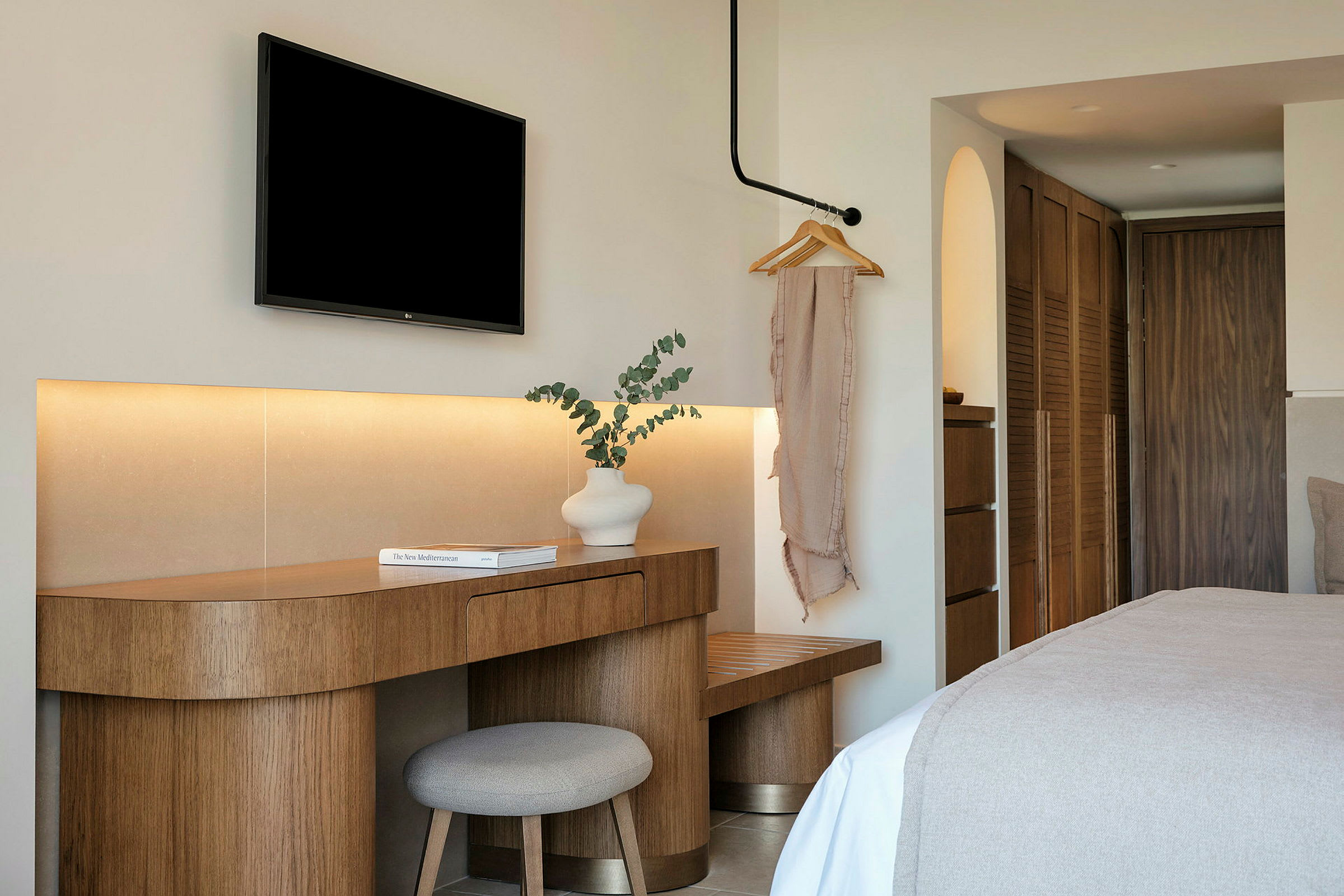 Ett hotellrum med en modern inredning. En träbord med en vit vas och en bok står bredvid en stol. En svart TV är monterad på väggen ovanför. (AI-genererad text)