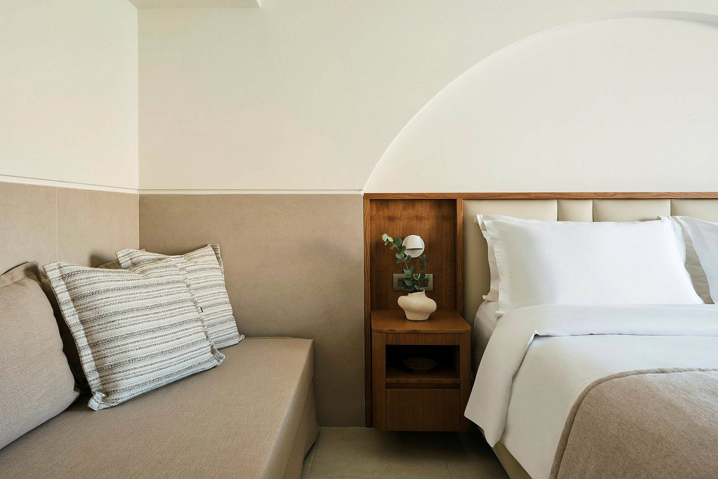 En ljus och modern hotellmiljö med en bäddad säng och en sittmöbel med kuddar. (AI-genererad text)