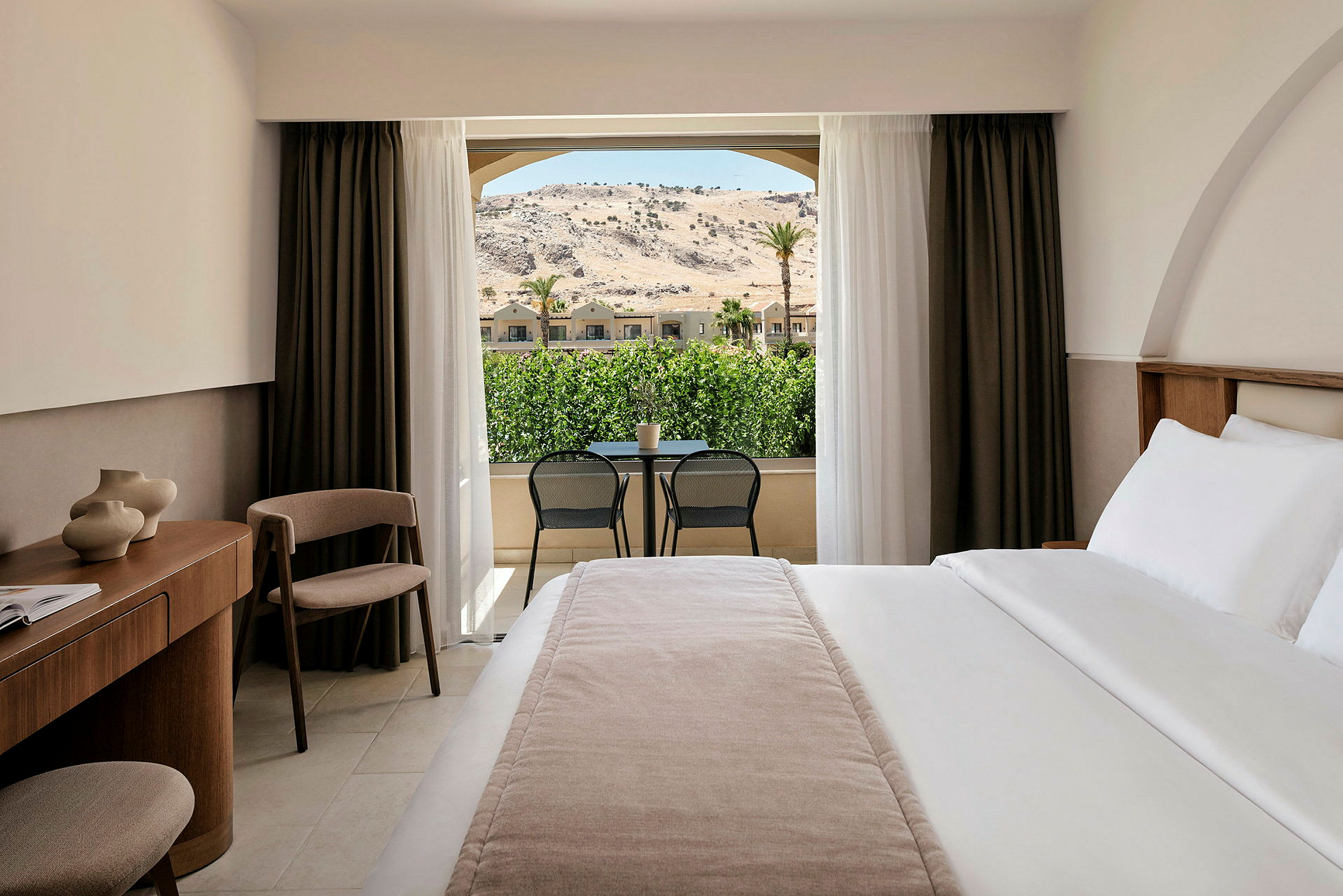 Hotellrum med en stor säng, skrivbord och stol. Utsikt mot grönska och berg genom glasdörrar. (AI-genererad text)