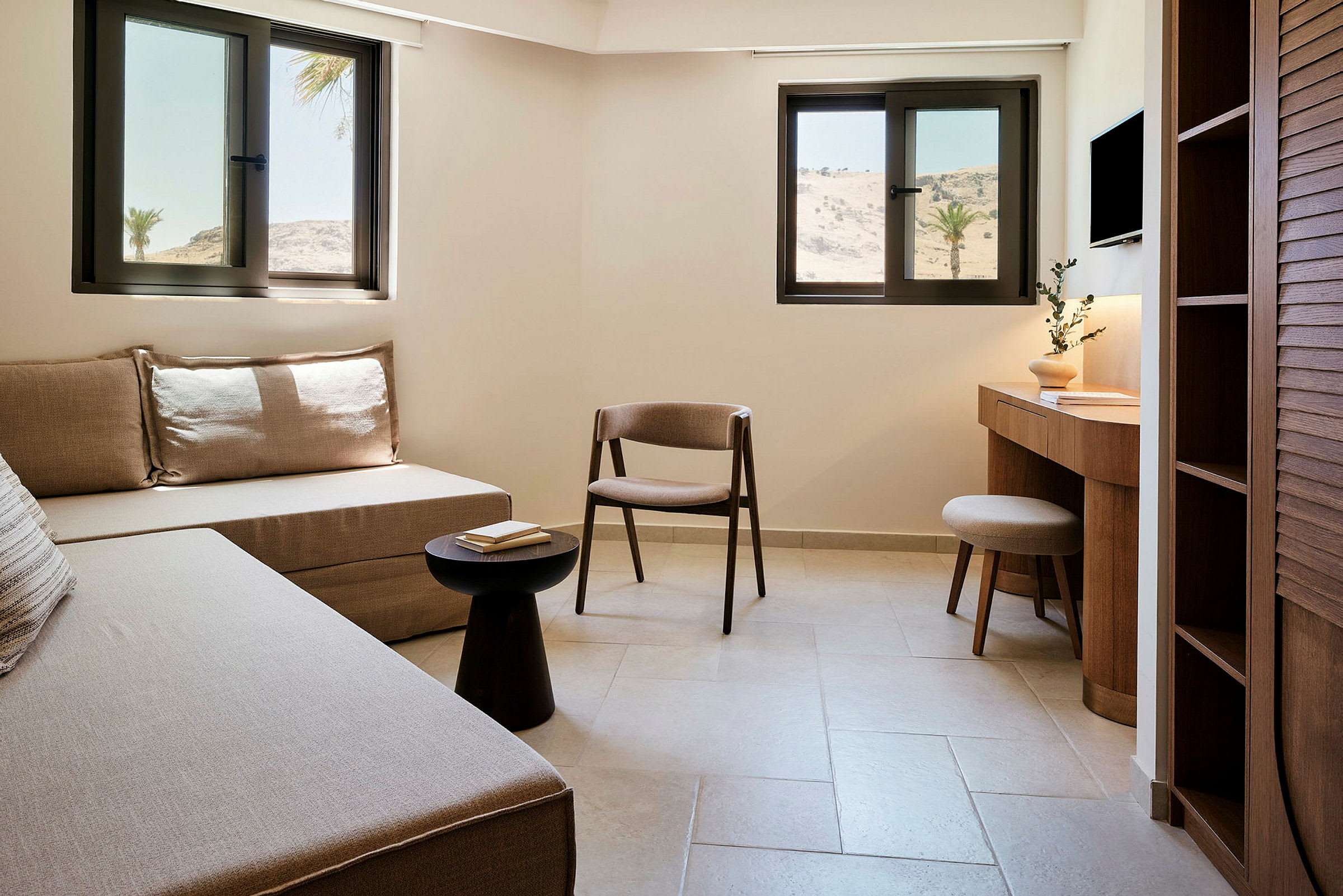 Ett hotellrum med soffa, stol, bord och skrivbord, samt fönster med utsikt över ett torrlagt landskap. (AI-genererad text)