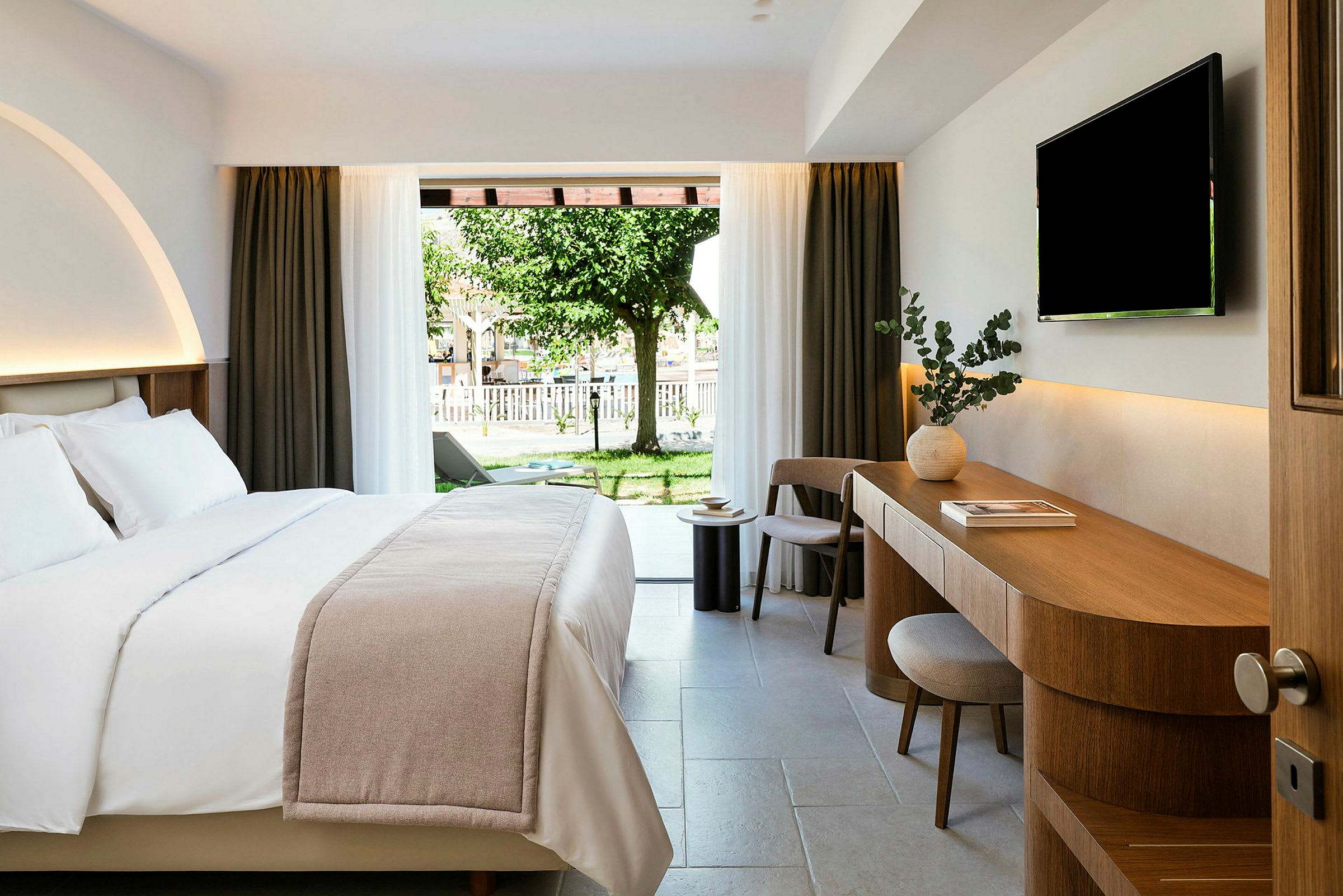 Ett hotellrum med en stor säng, en skrivbordsyta med stol och TV på väggen. Glasdörrar leder till en trädgård med gräs och ett träd. (AI-genererad text)