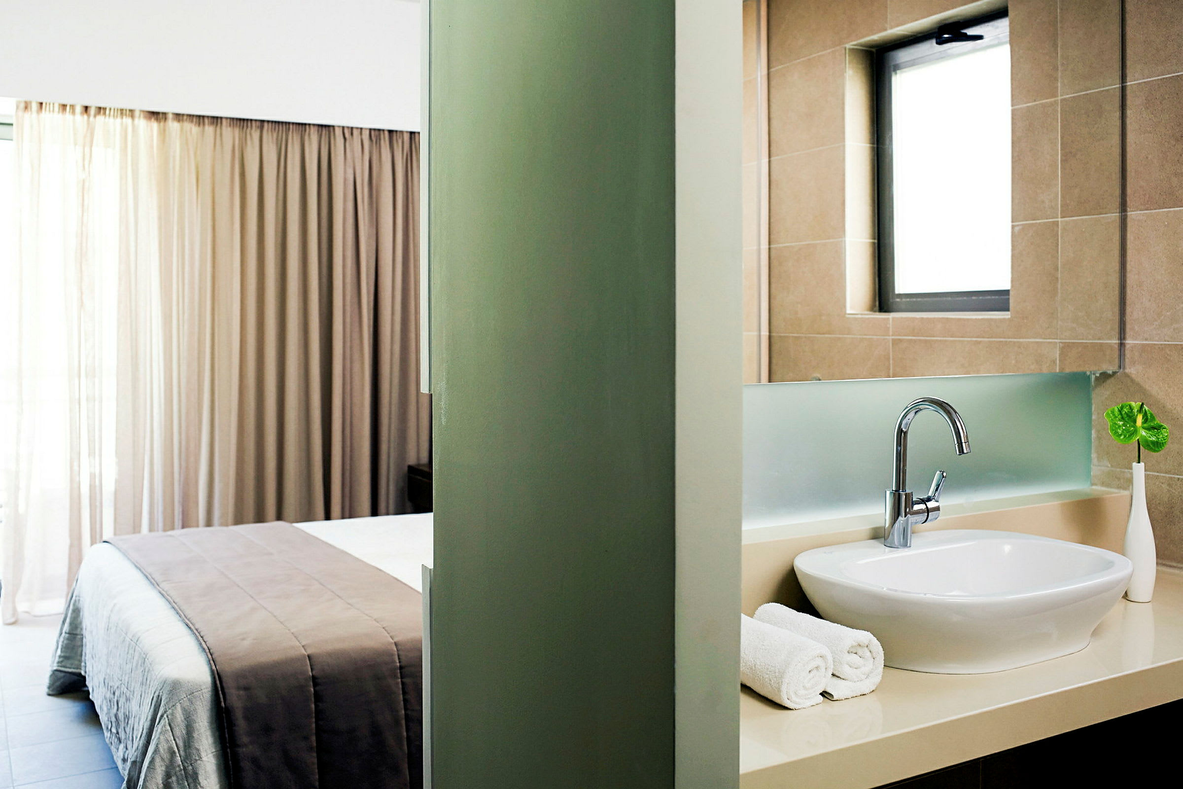 Ett hotellrum med en delad vy mellan sovrum och badrum. Sovrummet har en säng med beige sängkläder och en fönster med gardiner. Badrummet har en vit tvättställ, handdukar och ett litet grönt blad i en vas. (AI-genererad text)