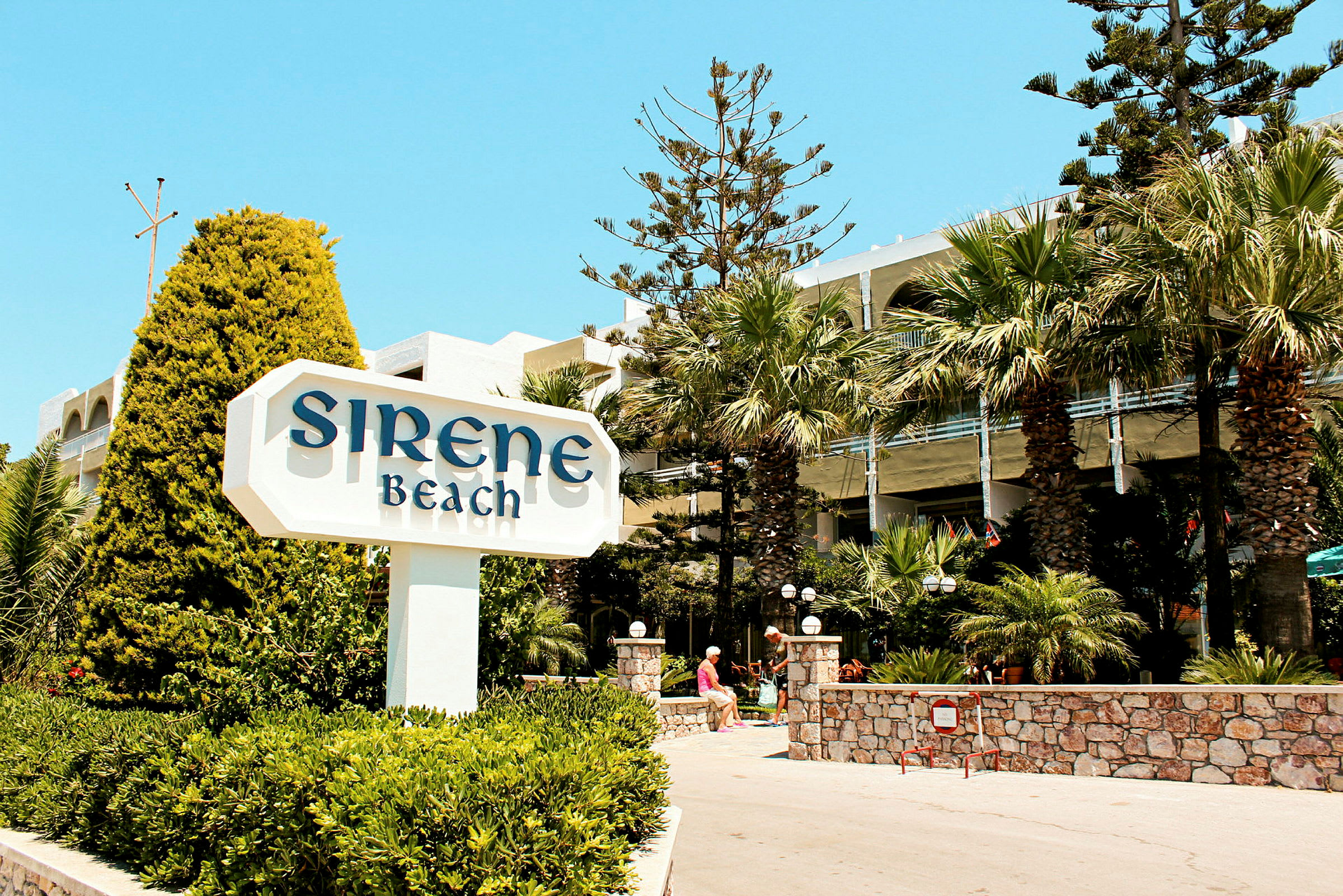 Skylt med texten 'Sirene Beach' framför ett hotell med palmer och grönska. (AI-genererad text)