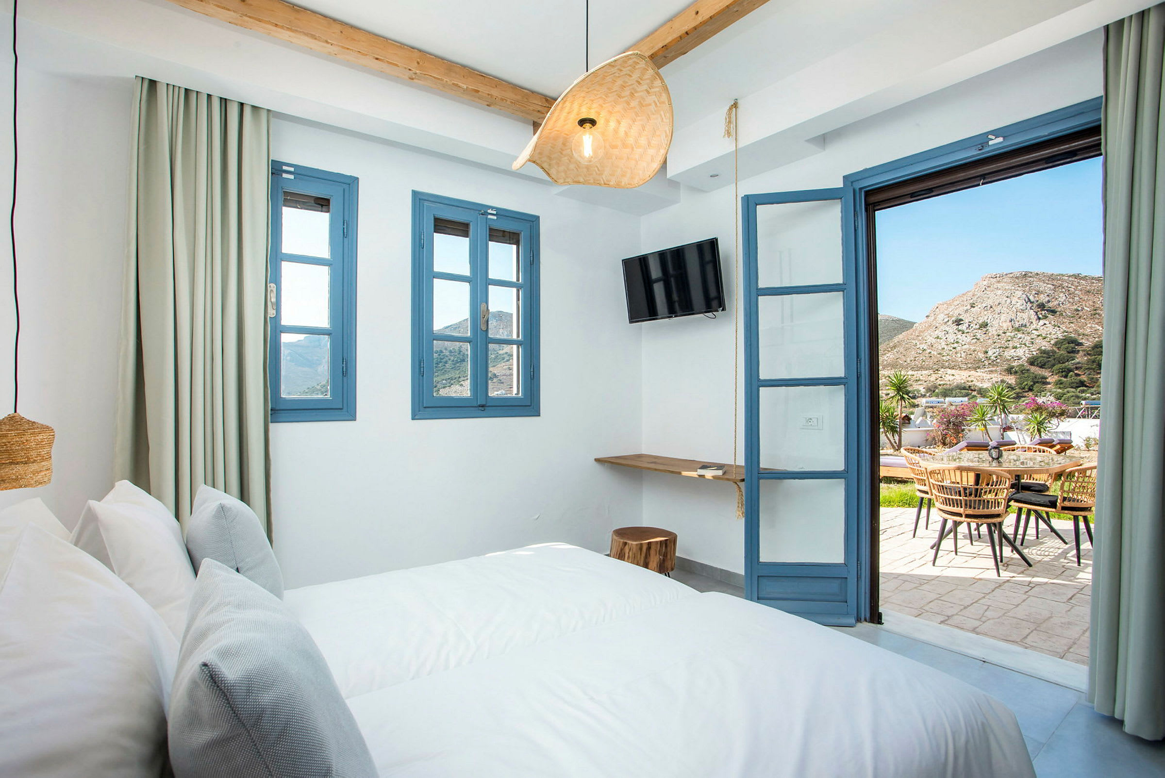 Ett hotellrum med säng, blå fönsterkarmar och dörr som öppnar mot en terrass med bord och stolar. (AI-genererad text)