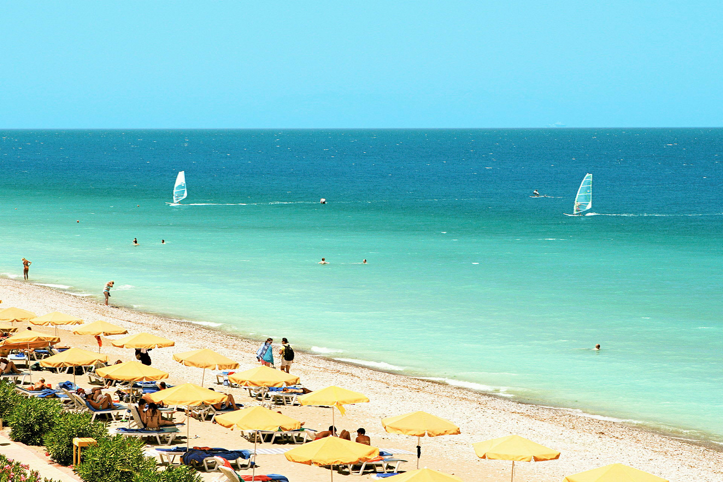 Strand med gula parasoll och solsängar vid havet, med människor som badar och vindsurfar. (AI-genererad text)