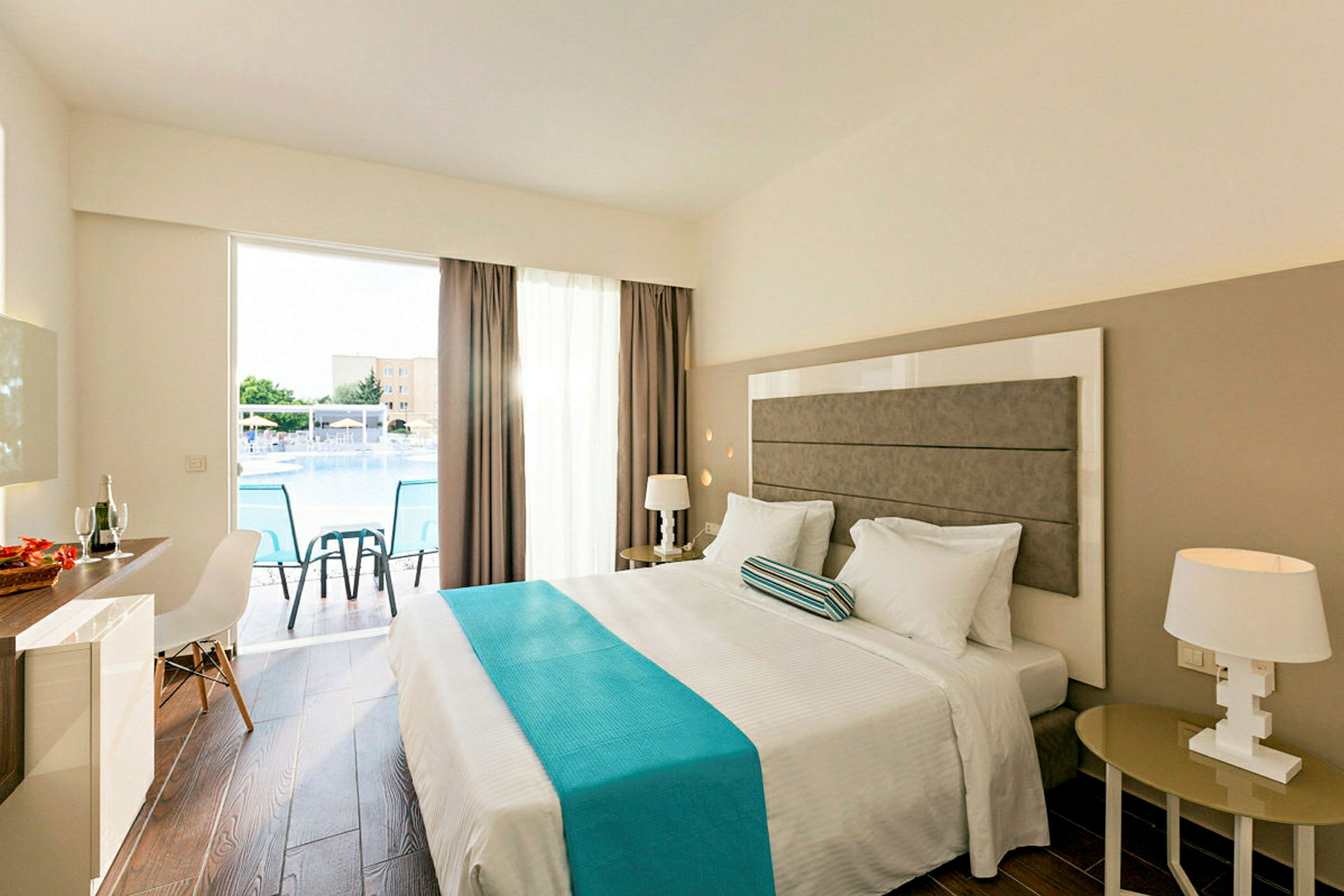 Ett hotellrum med en dubbelsäng, nattduksbord, en stol och ett skrivbord. Fönstret har utsikt över en pool. (AI-genererad text)