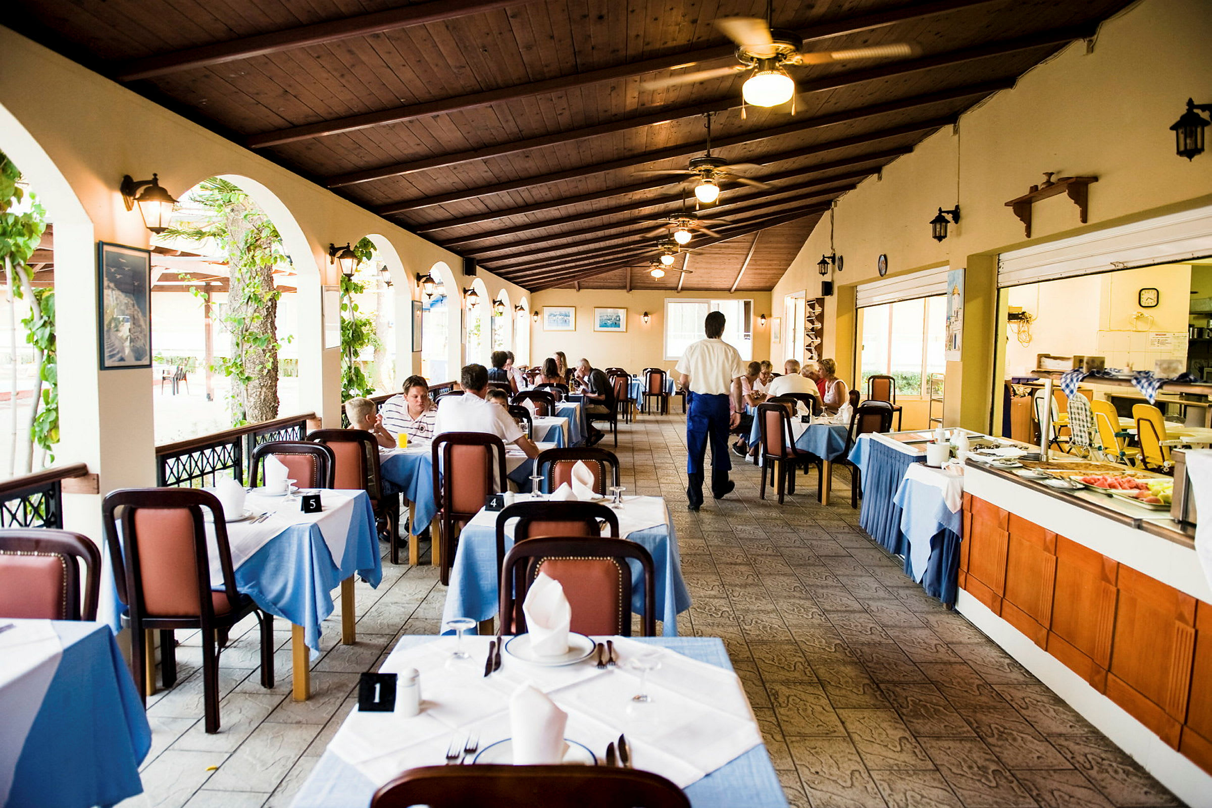 En restaurang med dukade bord och gäster som äter middag i en trevlig miljö. Fönsteröppningar med grönska syns på ena sidan. (AI-genererad text)