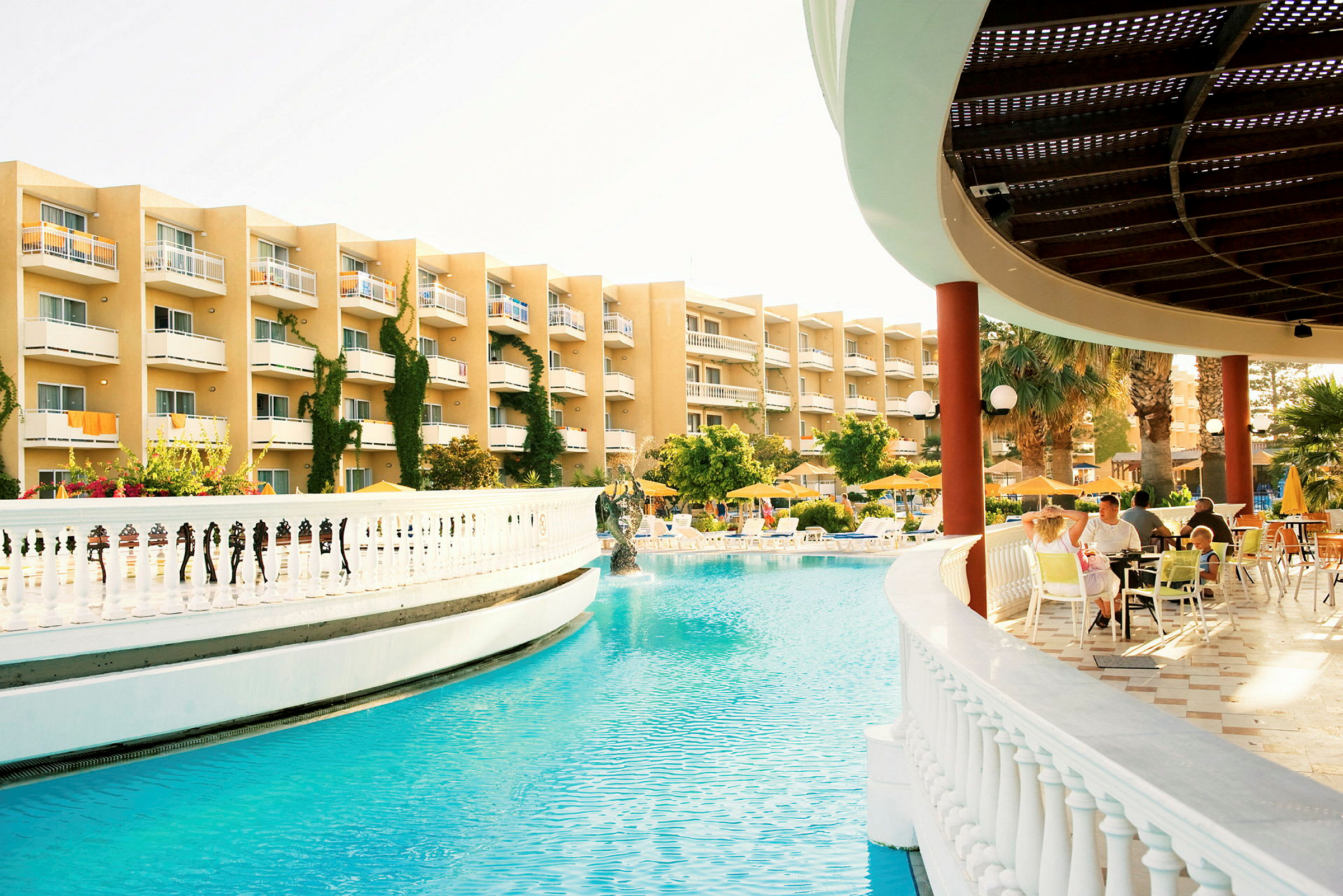En hotellpool omgiven av solsängar och parasoller, med en byggnad i bakgrunden och ett överbyggt område med sittplatser. (AI-genererad text)