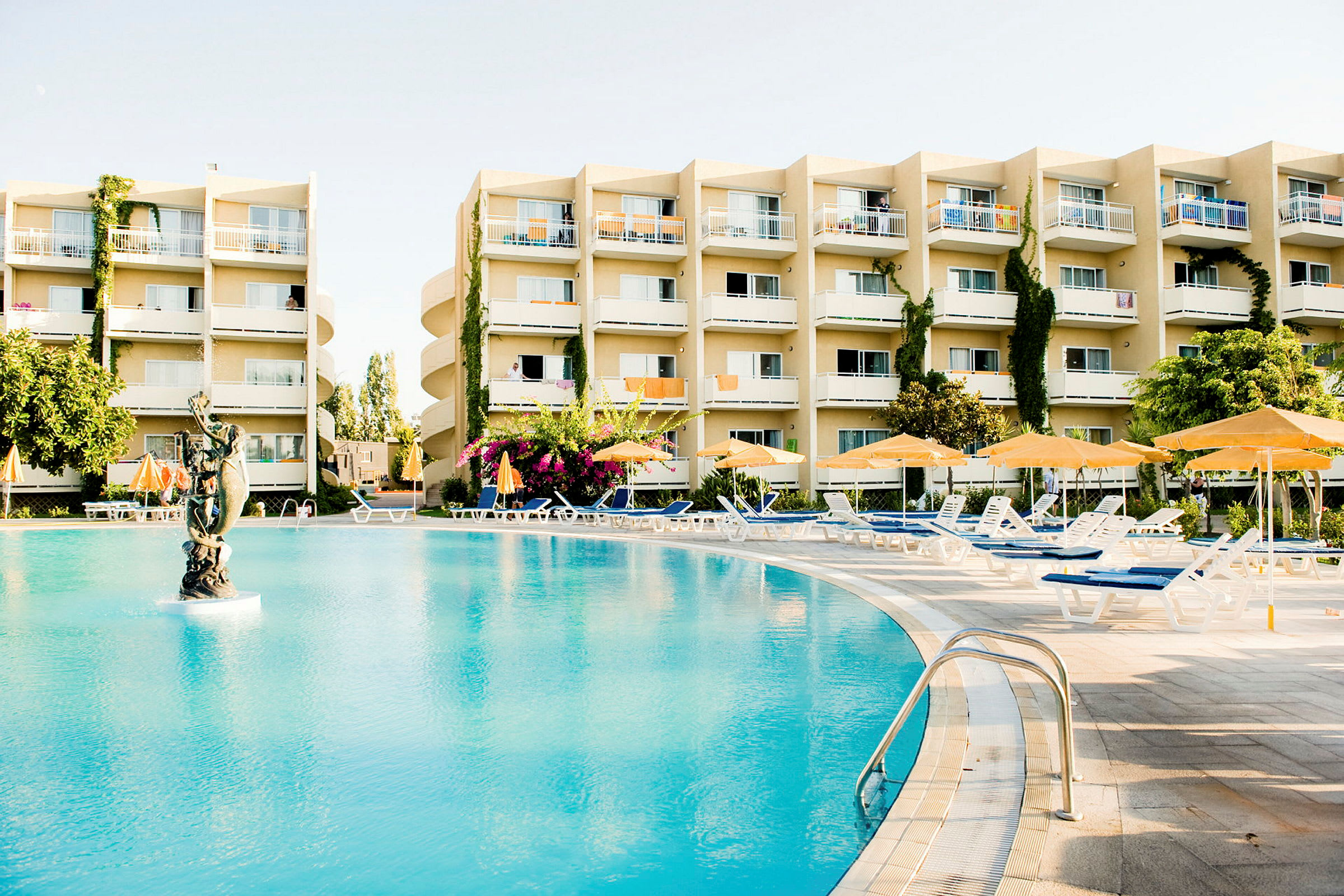 En utomhusbild föreställande en stor pool med en staty i mitten, omgiven av solstolar och parasoller framför en hotellbyggnad. (AI-genererad text)