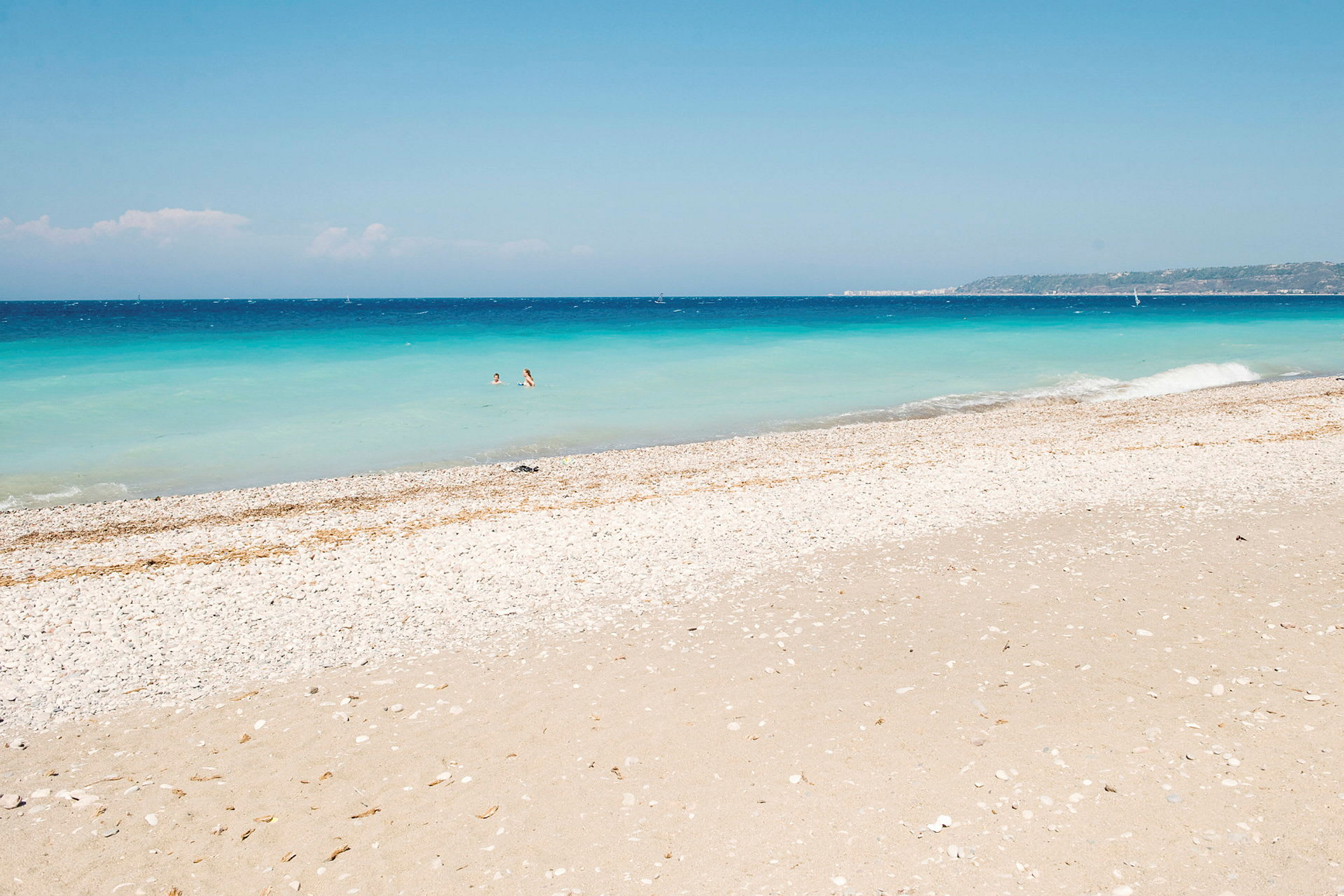 Vit klapperstensstrand vid havet med klart blått vatten och två personer som badar. (AI-genererad text)