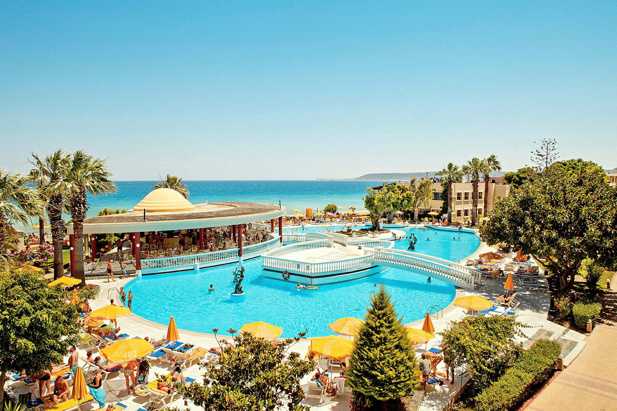 En semesterresort med poolområde, soldäck, parasoller och utsikt över kusten och havet i bakgrunden. (AI-genererad text)