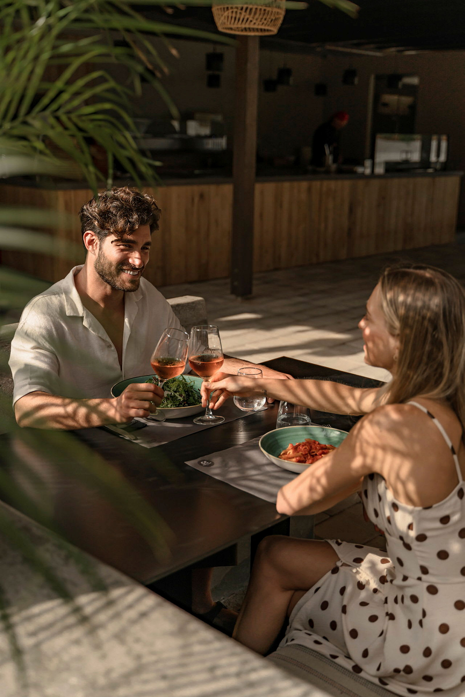 Två personer skålar med glas innehållande rosévin vid ett bord på en solig restaurang. (AI-genererad text)