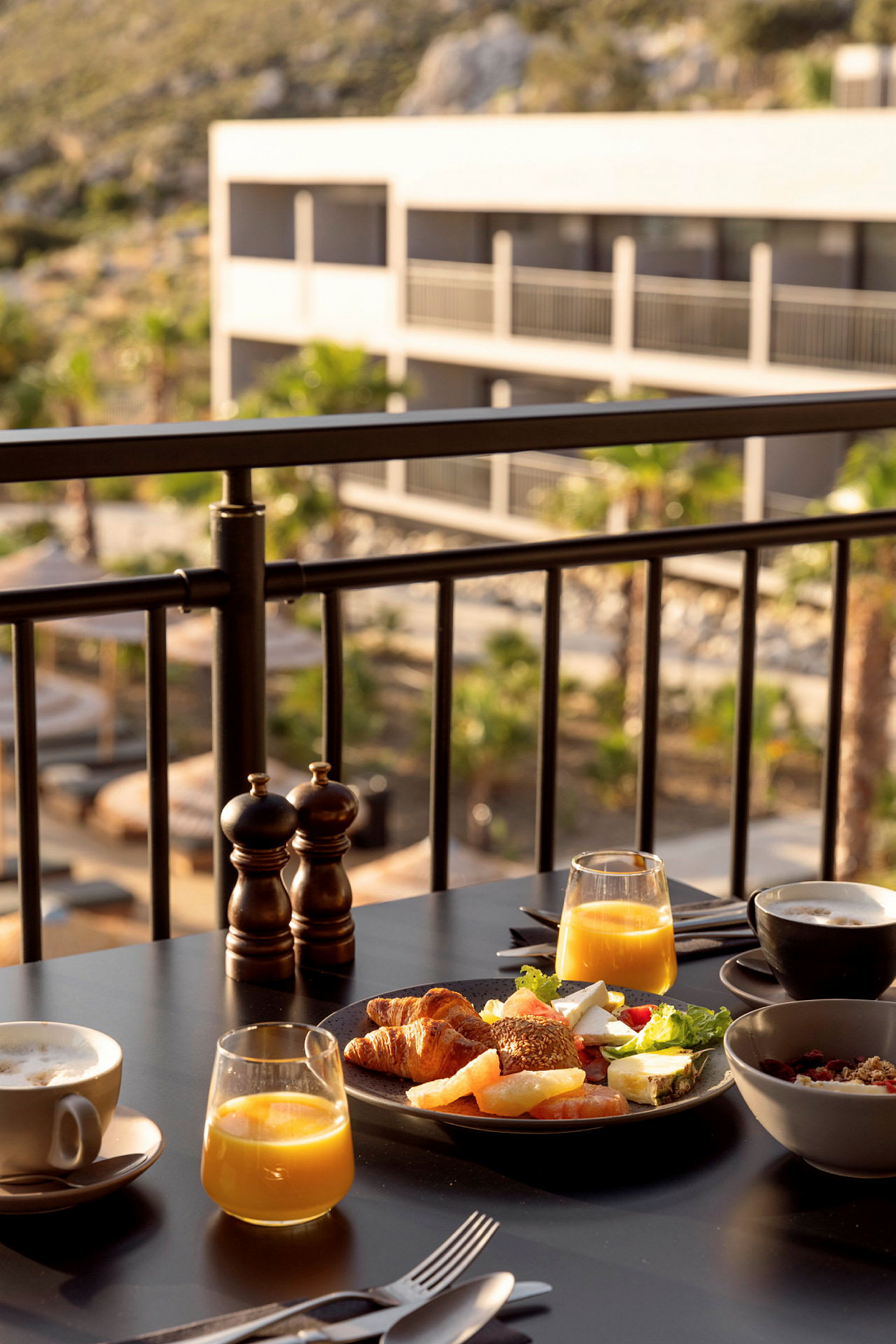 Frukost med juice, kaffe och olika bröd serverad vid ett bord på ett soldäck med utsikt över en hotellbyggnad. (AI-genererad text)