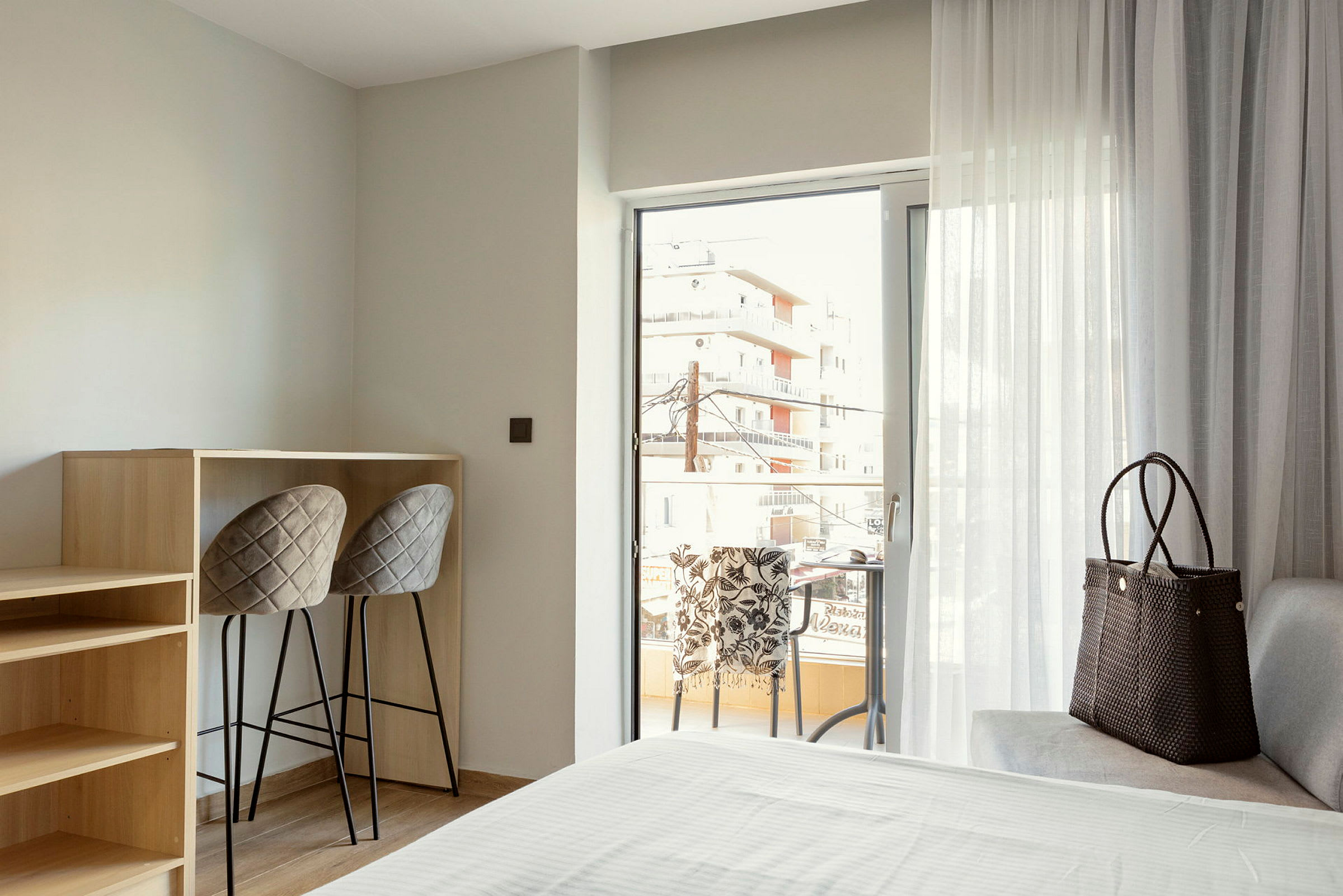 Ett hotellrum med balkong, två stolar vid en hög bänk, och en soffa med en väska placerad ovanpå. (AI-genererad text)