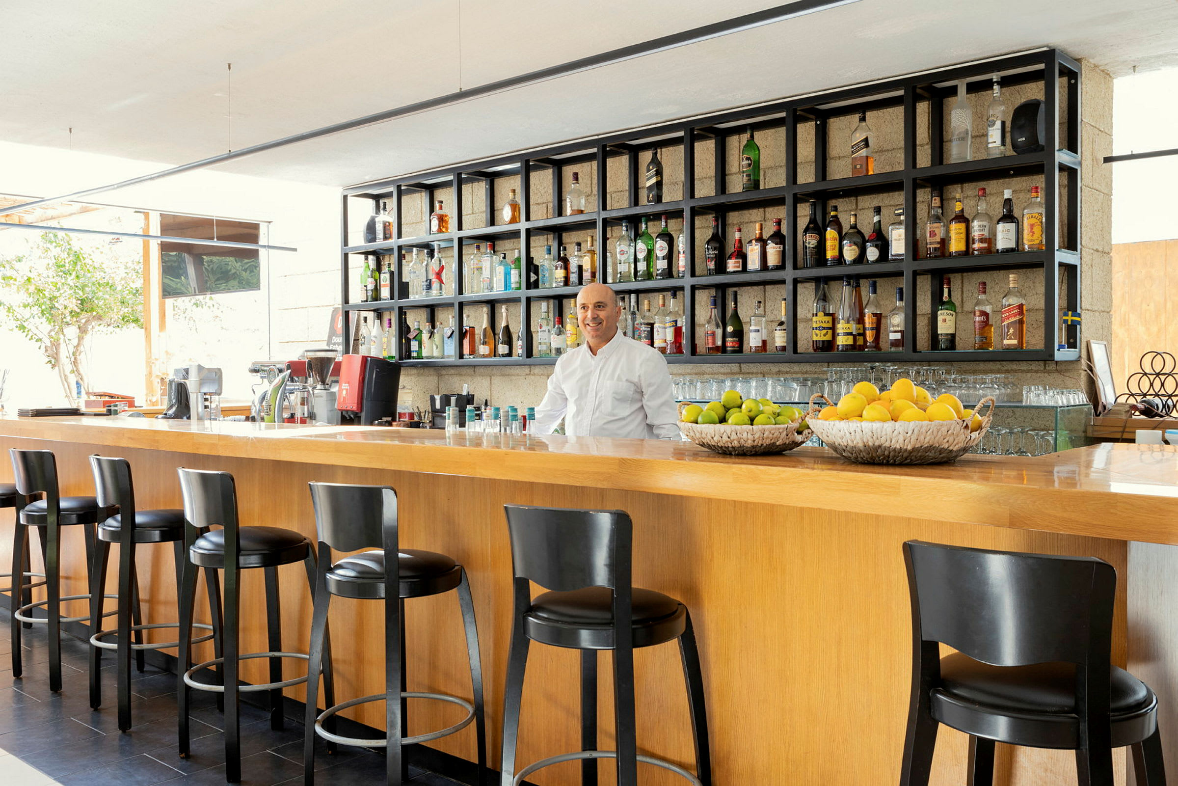 En modern bar med höga stolar vid en trädisk, hyllor fyllda med flaskor och fruktkorgar med citrusfrukter. (AI-genererad text)