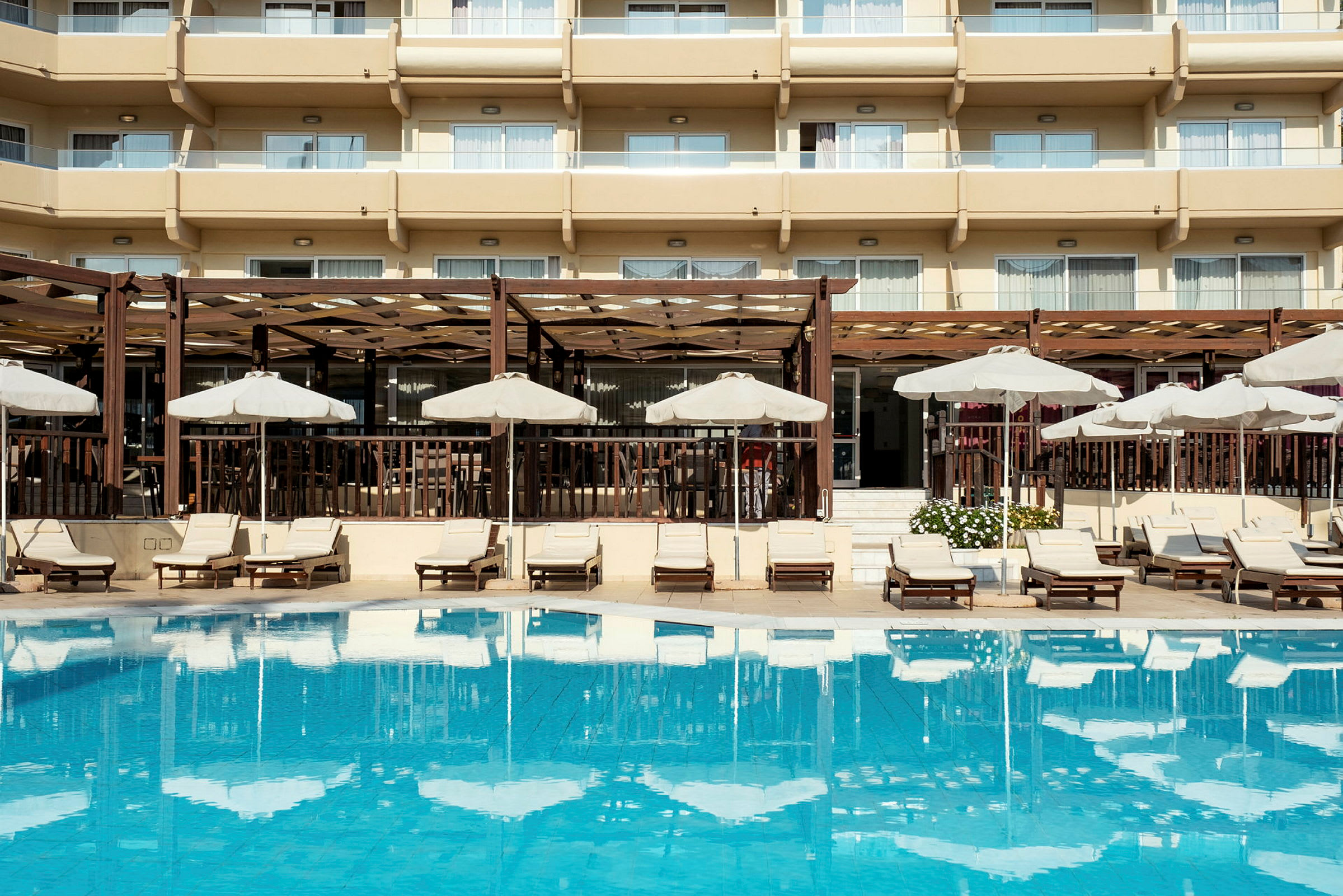 Vy över en pool med solstolar och parasoller framför en byggnad med balkonger. (AI-genererad text)