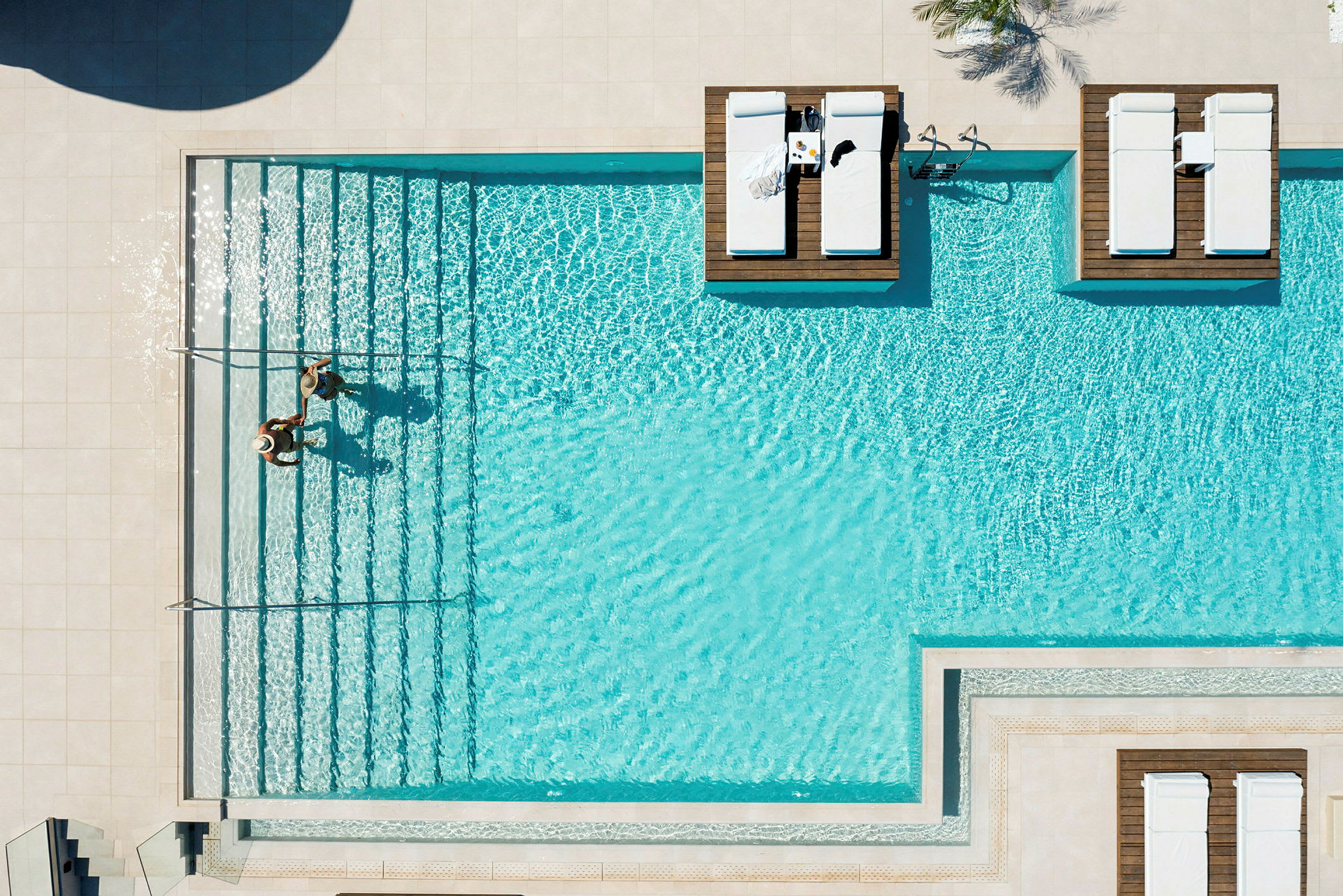 Vy ovanifrån av en pool med solsängar på träplattformar och två personer som står vid pooltrappan. (AI-genererad text)
