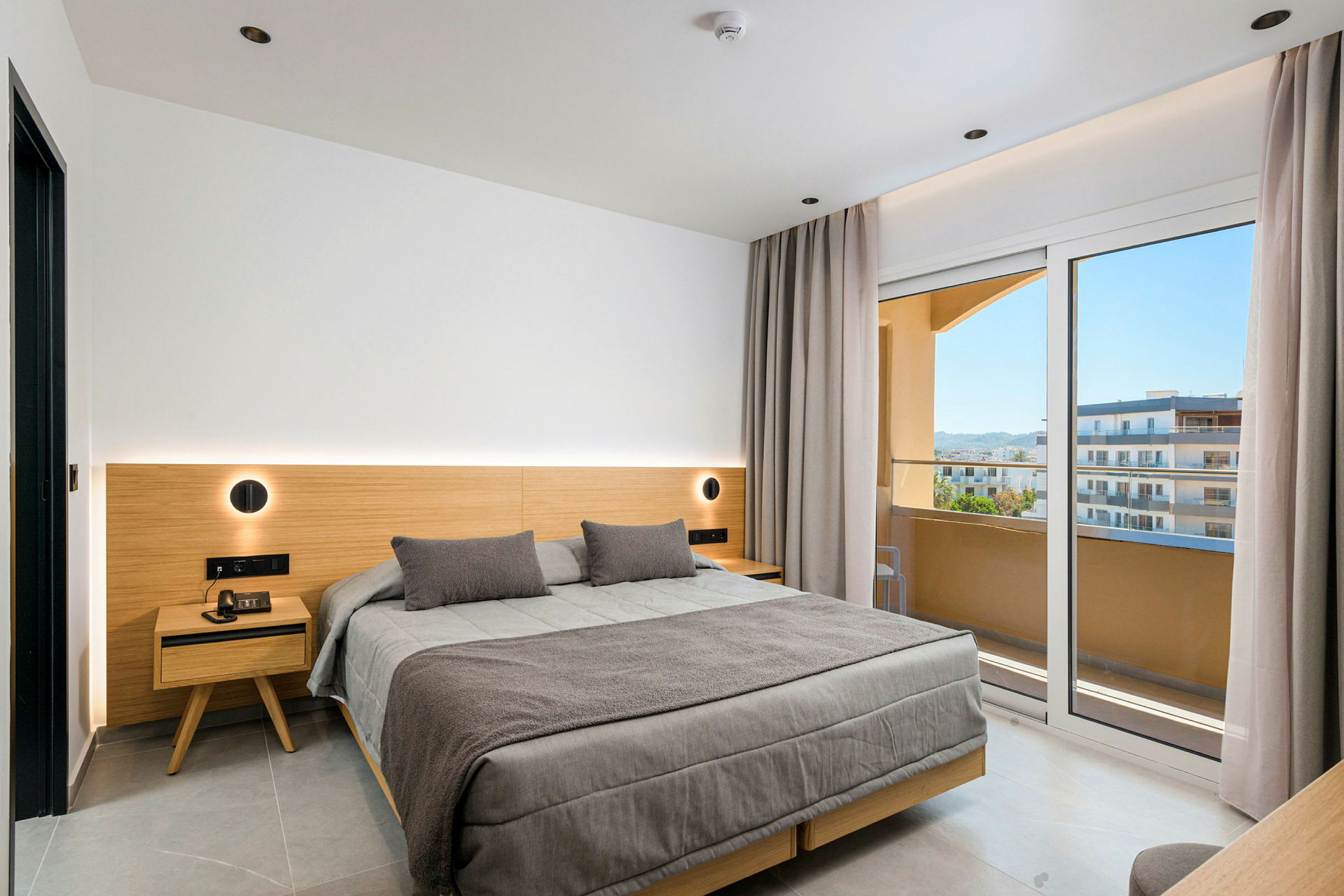 Ett modernt hotellrum med stor säng, trädetaljer och glasdörrar som leder ut till en balkong med utsikt. (AI-genererad text)