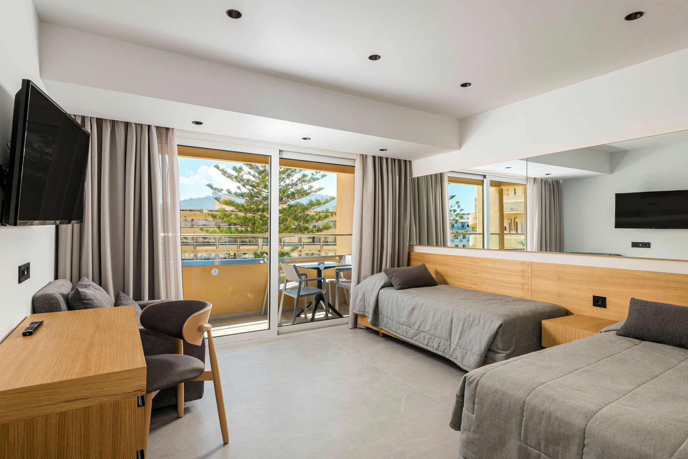 Ett hotellrum med två enkelsängar, en skrivbordsmöbel, en stol, en väggmonterad TV och en balkong med möbler samt utsikt över området utanför. (AI-genererad text)
