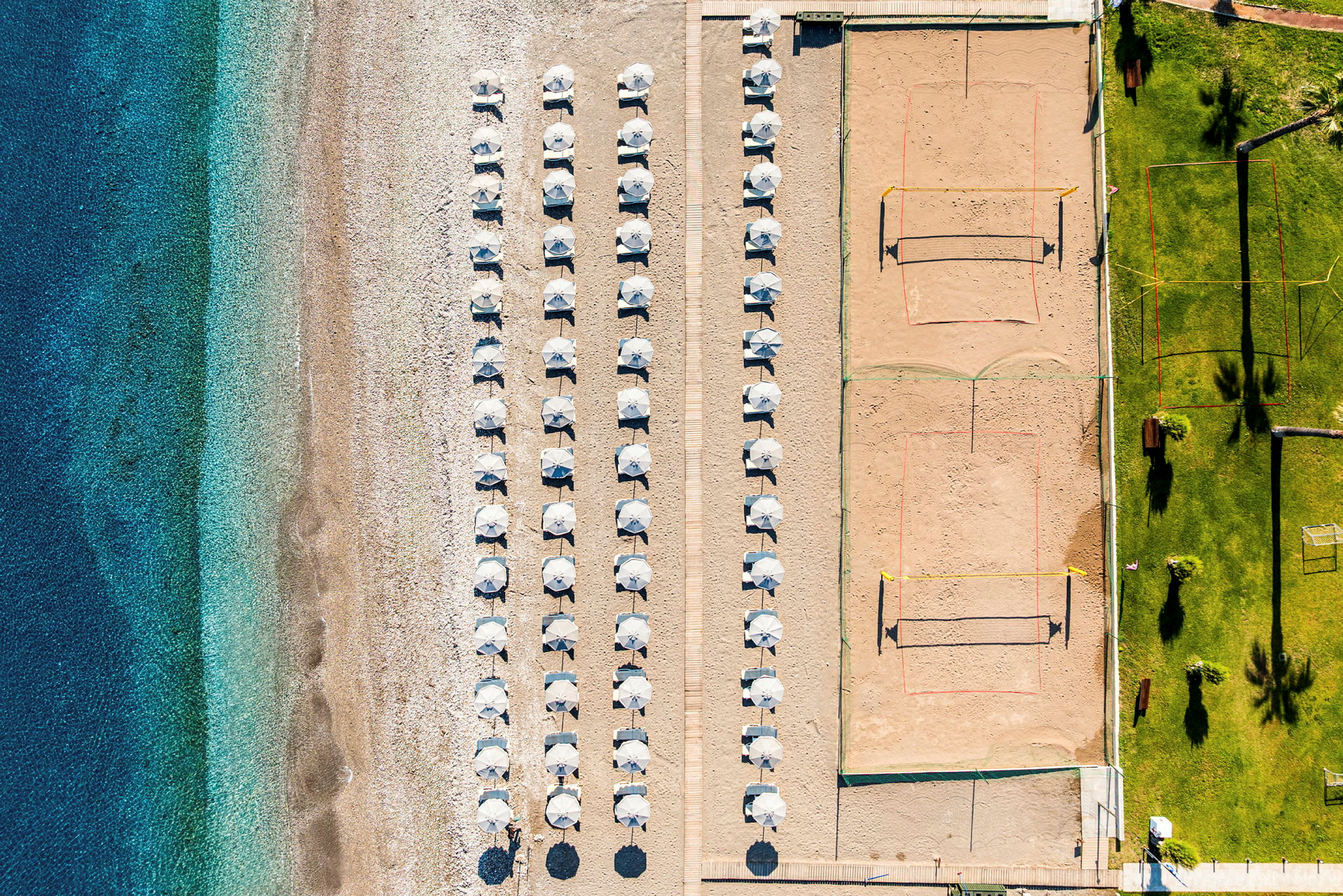 Flygfoto av en strand med parasoller uppställda i rader, en volleybollplan på sand vid sidan och en gräsmatta med palmer och sittplatser. (AI-genererad text)