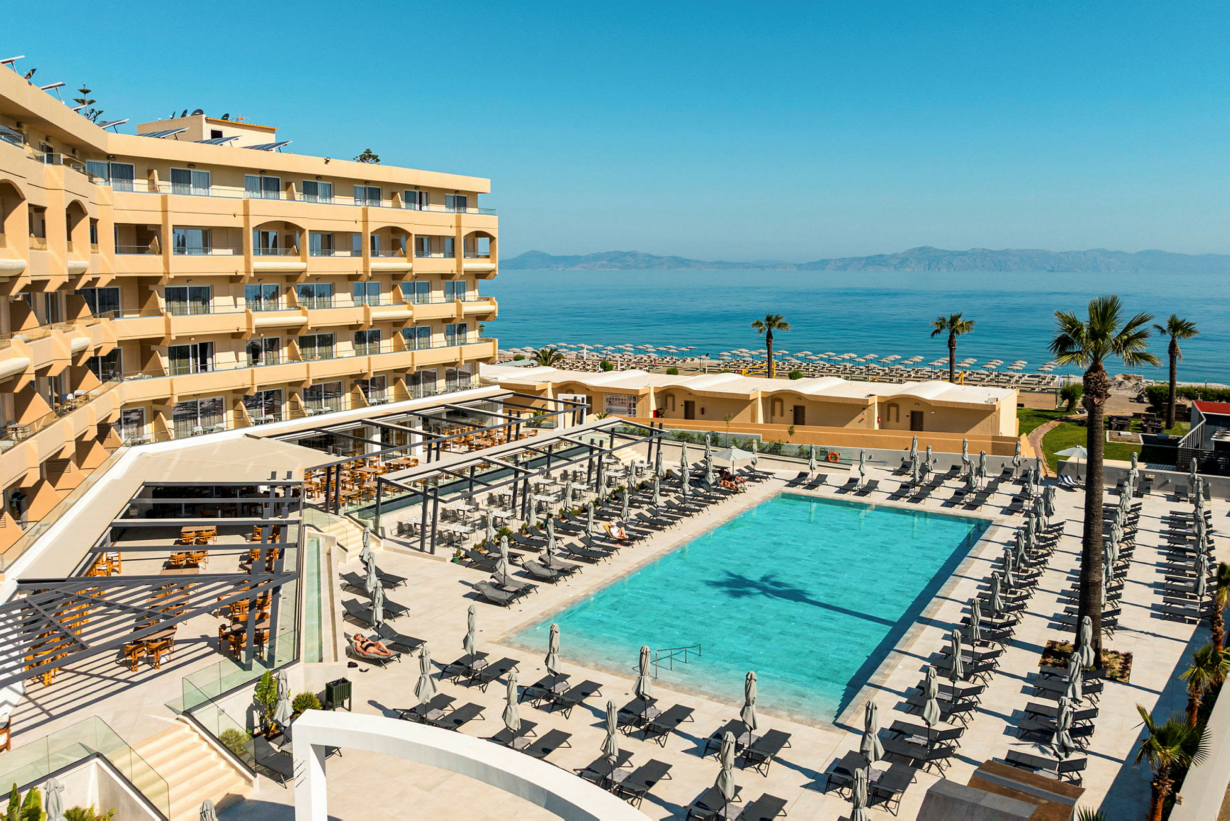 Ett hotellområde med en stor pool, omgiven av solstolar och parasoller. I bakgrunden syns stranden och havet. (AI-genererad text)