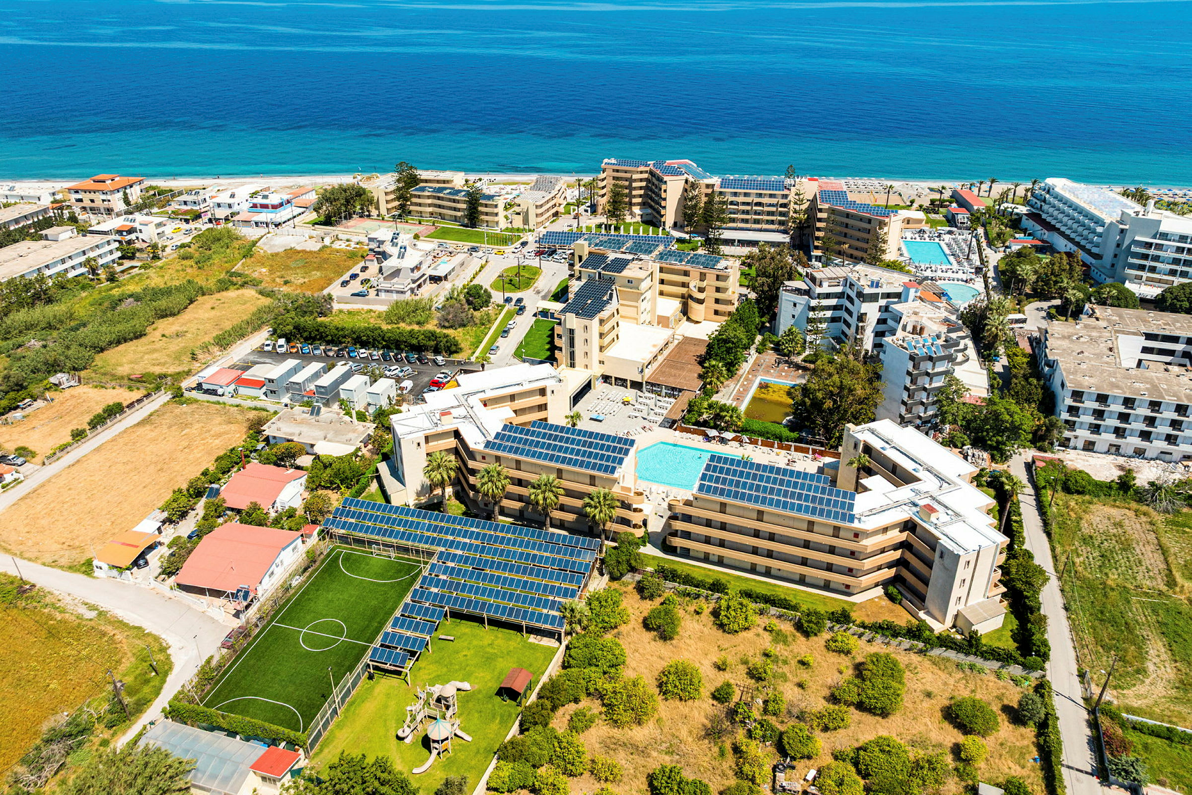 Flygfoto över ett resortområde med hotell, pooler, grönområden och en lekplats, beläget nära stranden och havet. (AI-genererad text)
