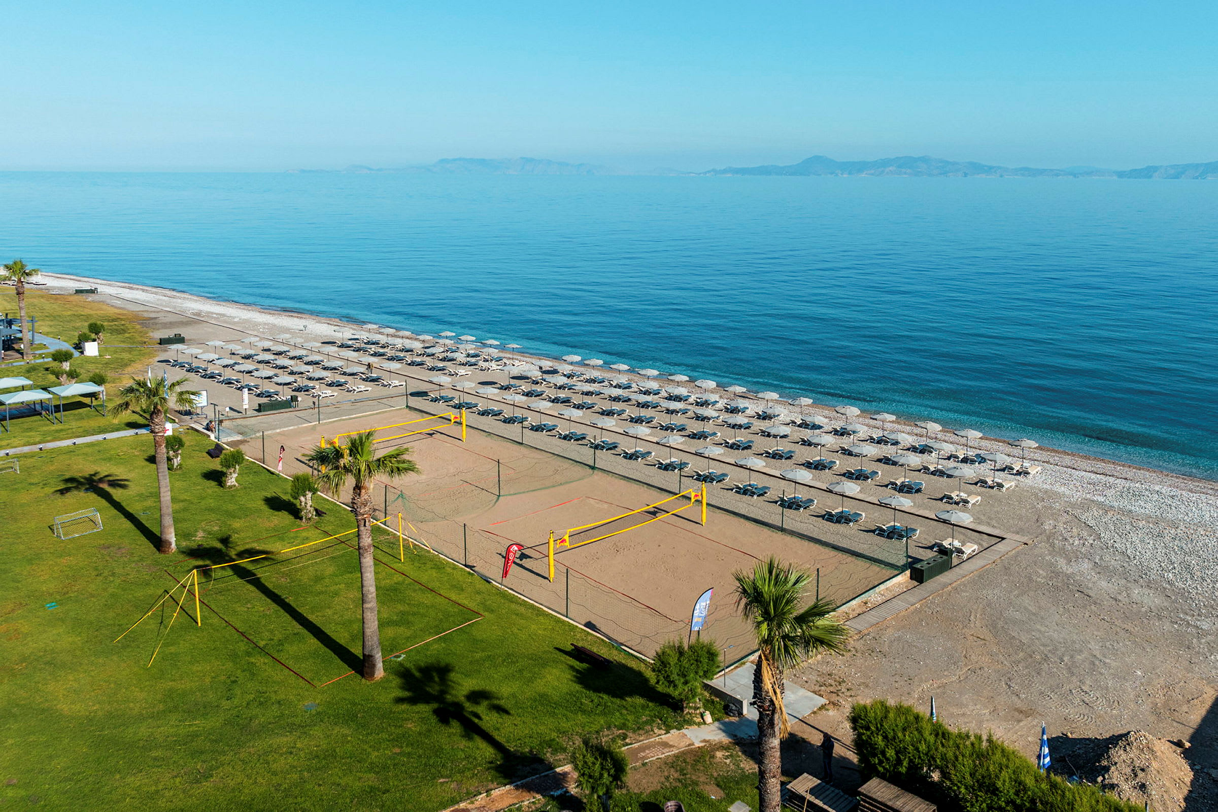 Bild av en strand med solstolar och parasoller i flera rader, en sandstrand och ett volleybollnät bredvid ett grönområde. (AI-genererad text)