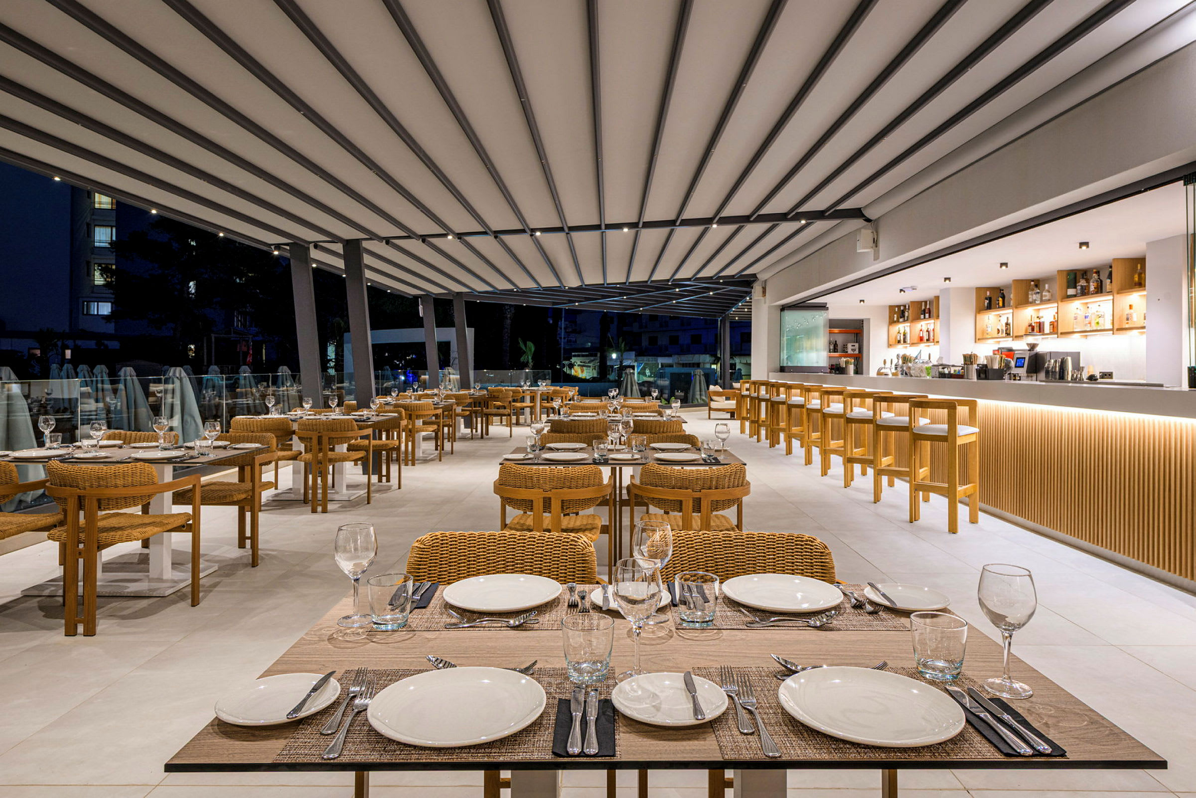 En modern restaurang med dukade bord och stolar i rotting samt en lång bardisk i bakgrunden. (AI-genererad text)