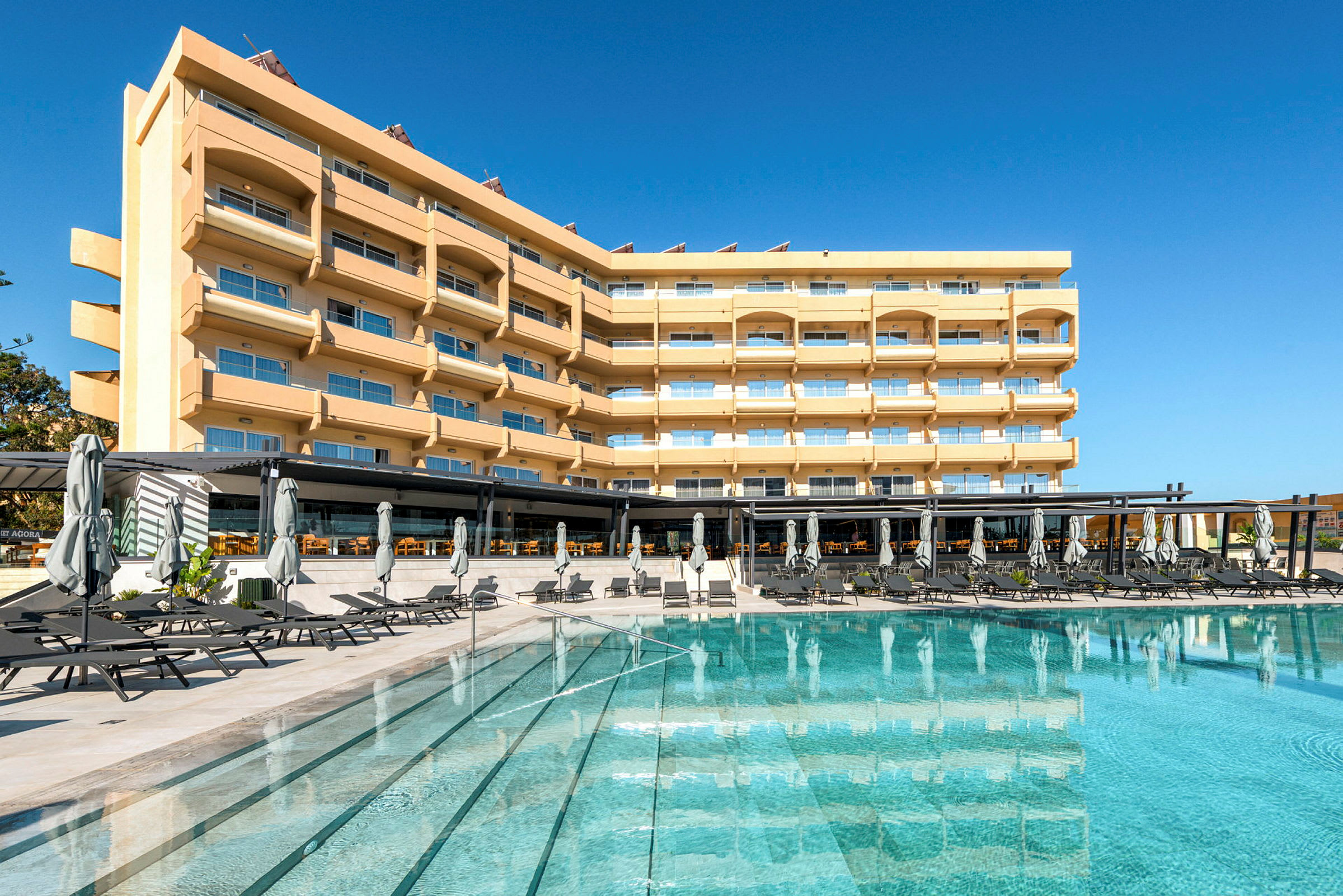 En hotellbyggnad med flera våningar, balkonger och stora fönster, framför en pool med solstolar och parasoller. (AI-genererad text)