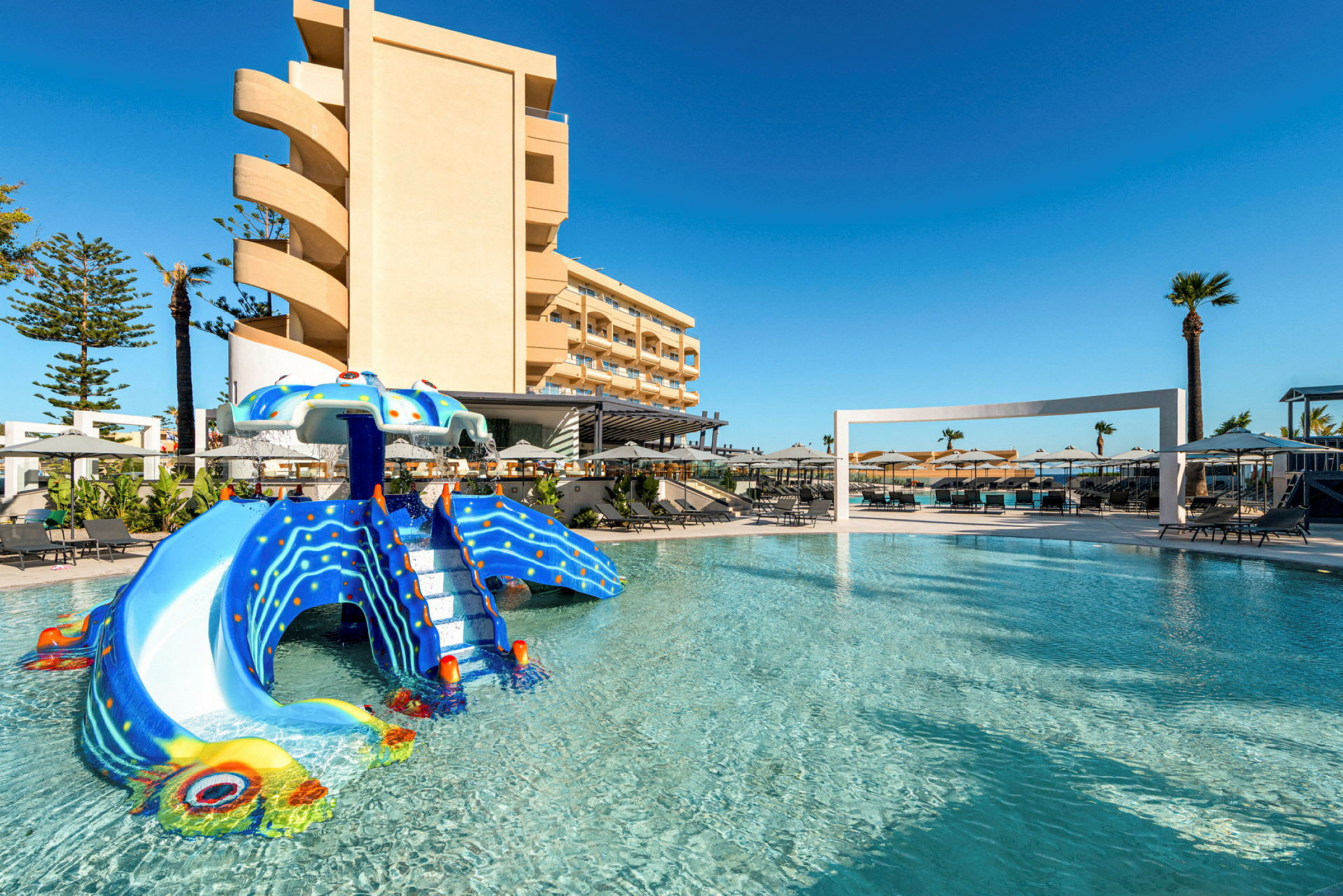 Bild av en pool med en lekfull rutschkana formad som en bläckfisk, omgiven av solstolar och ett hotell i bakgrunden. (AI-genererad text)