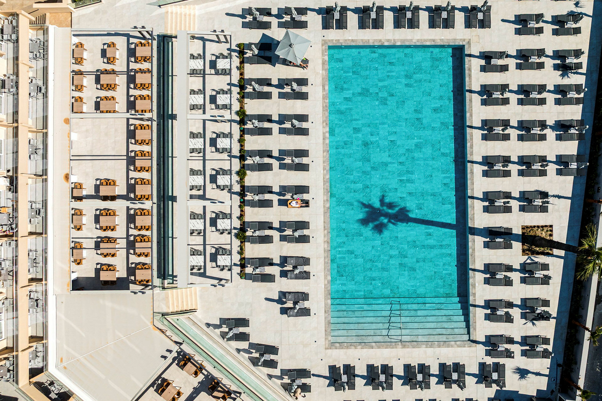Flygfoto över en stor, rektangulär pool omgiven av solstolar och parasoller. (AI-genererad text)