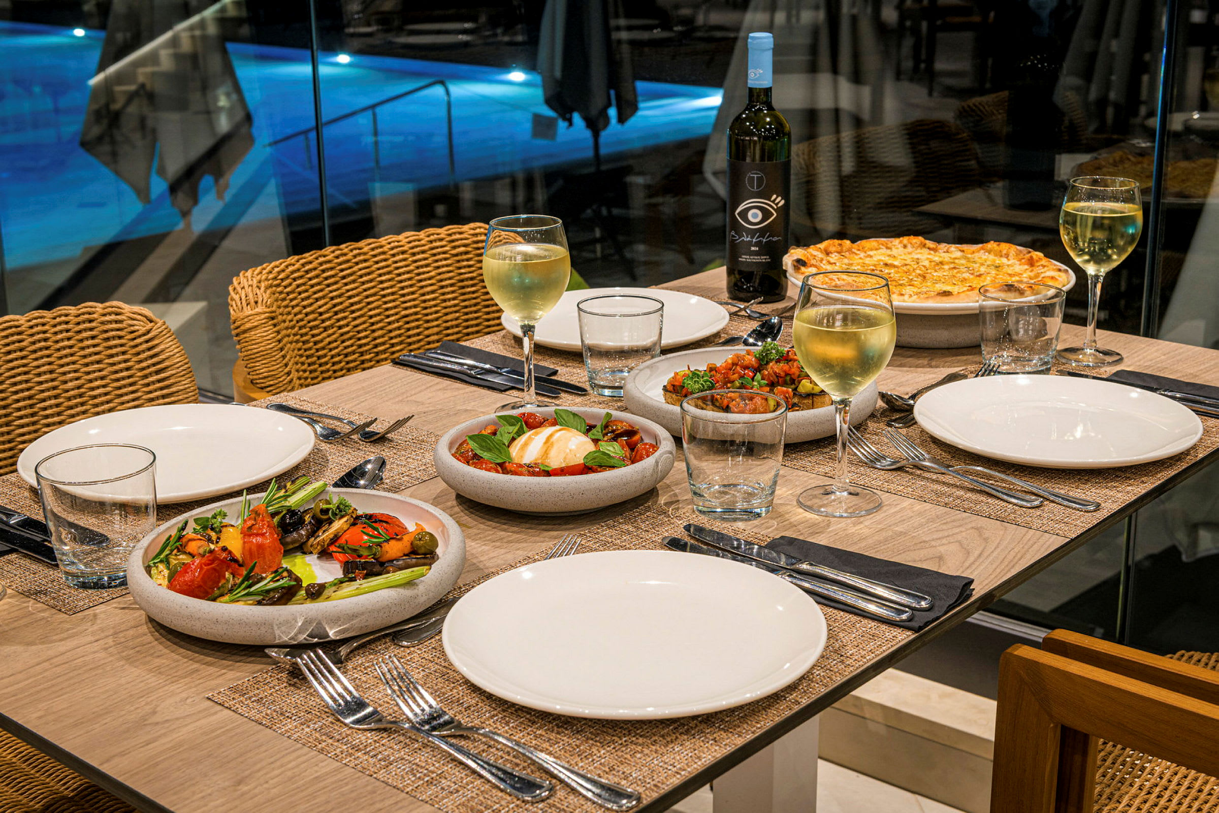 Ett dukat bord med tallrikar, vinglas, flaskor och olika maträtter. En bakgrund med en upplyst pool. (AI-genererad text)