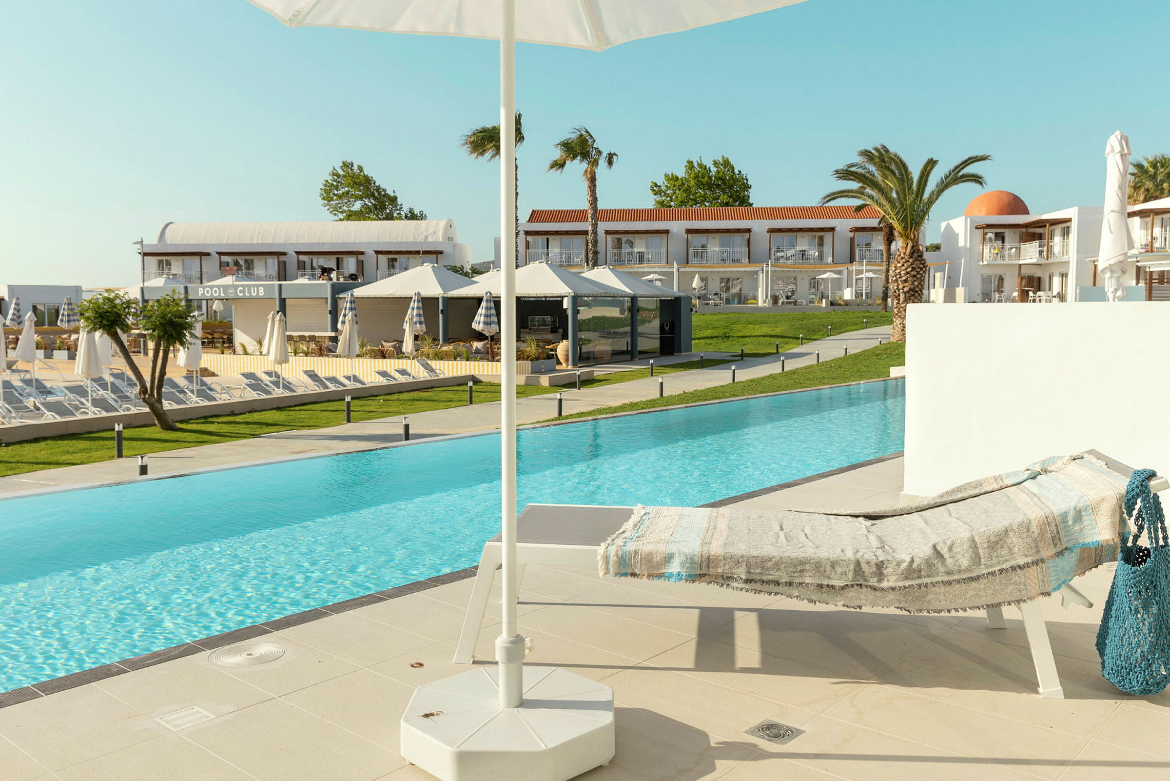 En solstol med en filt vid kanten av en pool, med en parasoll i förgrunden och hotellbyggnader i bakgrunden. (AI-genererad text)