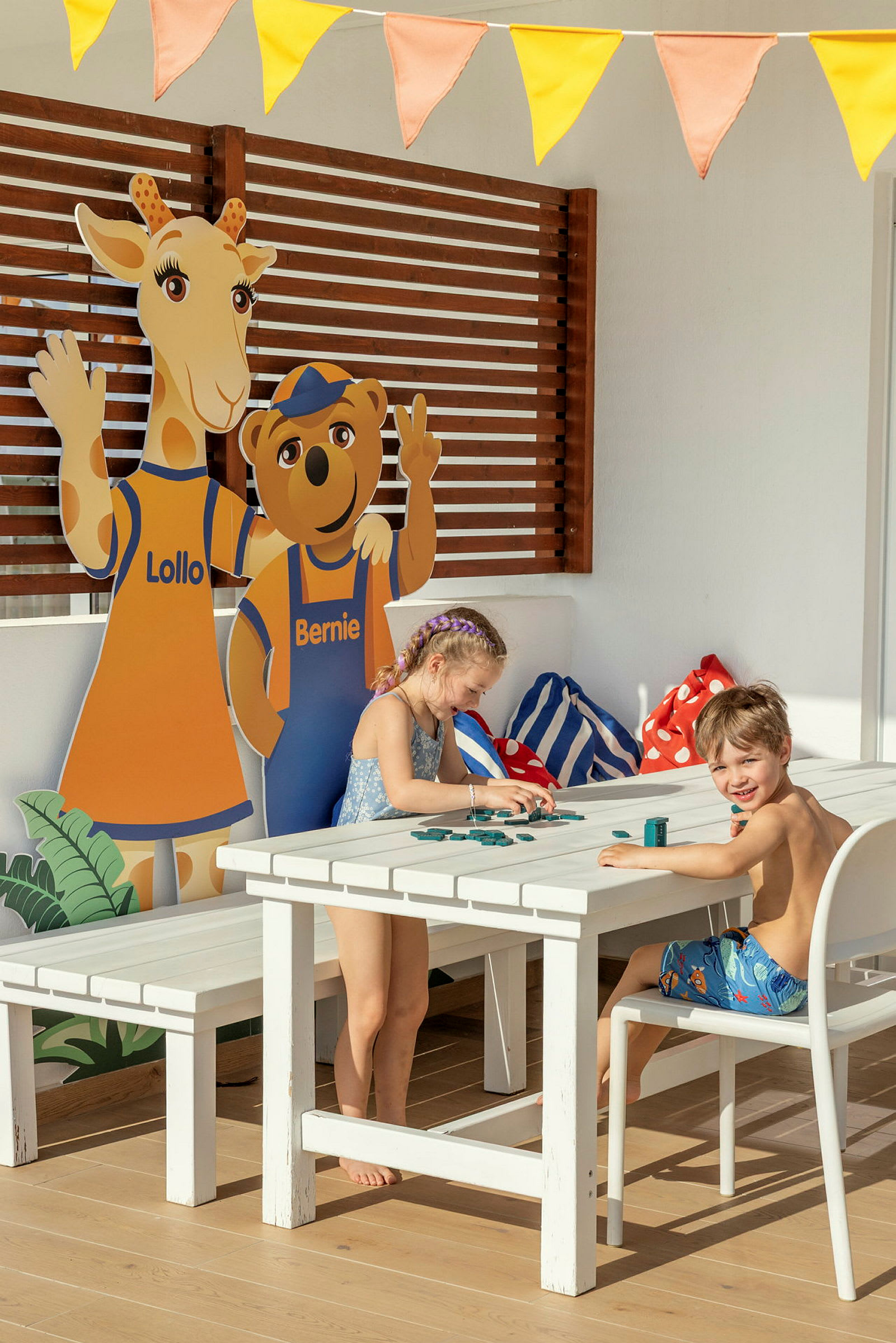 Två barn sitter vid ett utomhusbord och leker med spelbrickor. I bakgrunden syns cutouts av figurerna Lollo och Bernie. (AI-genererad text)