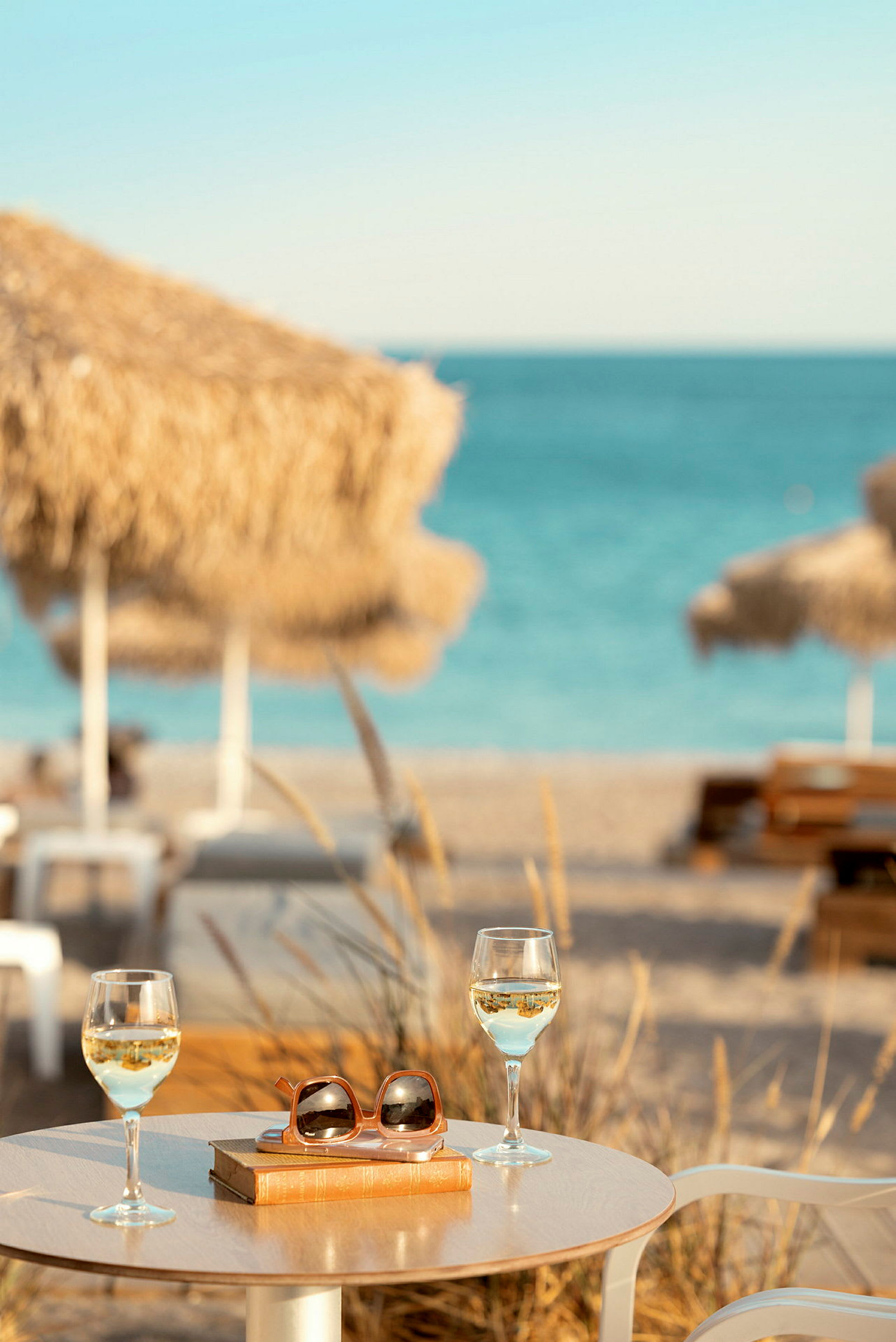 Bord med två glas vitt vin, en bok och solglasögon vid strand med parasoller och hav i bakgrunden. (AI-genererad text)